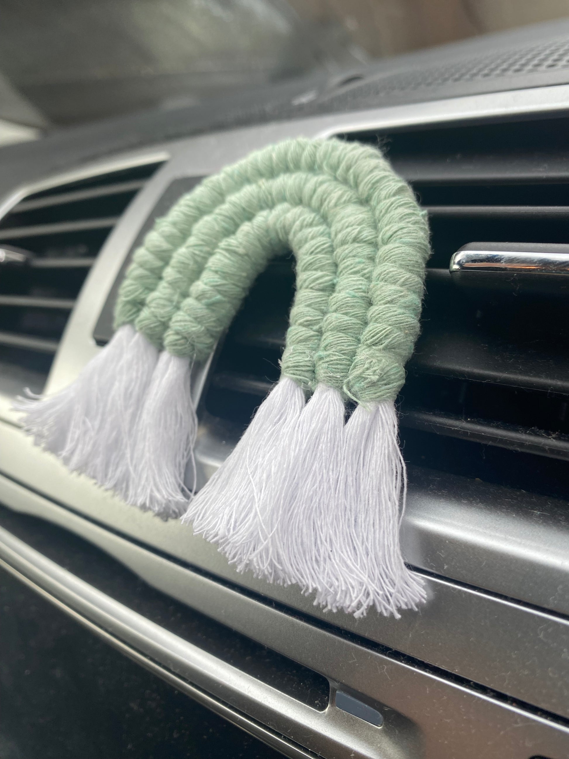 A mini macrame rainbow in green, attached to the car air vent on a black car interior.