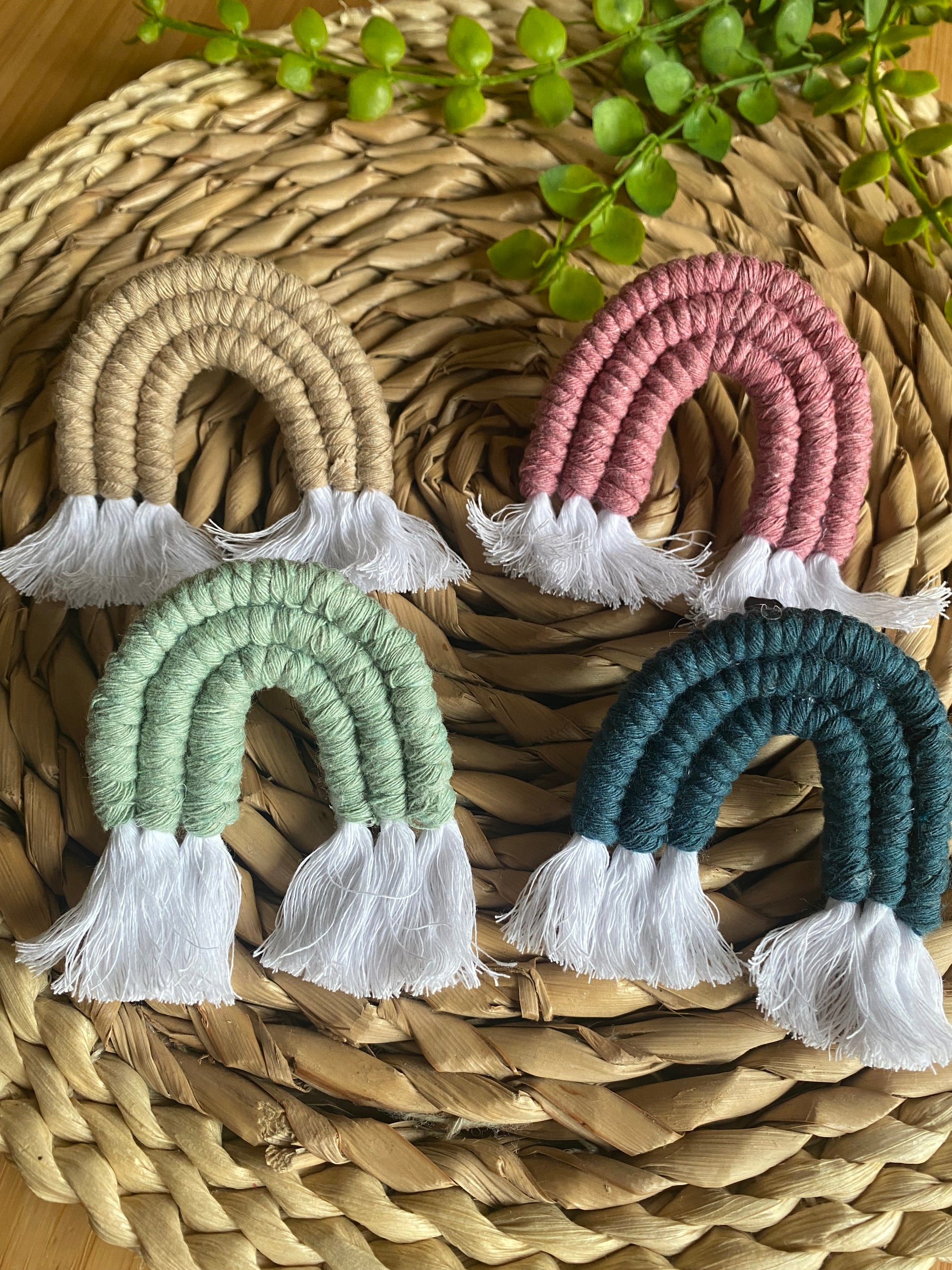 A set of four mini macrame rainbows in beige, pink, green and blue, laid next to each other on a woven wicker mat.