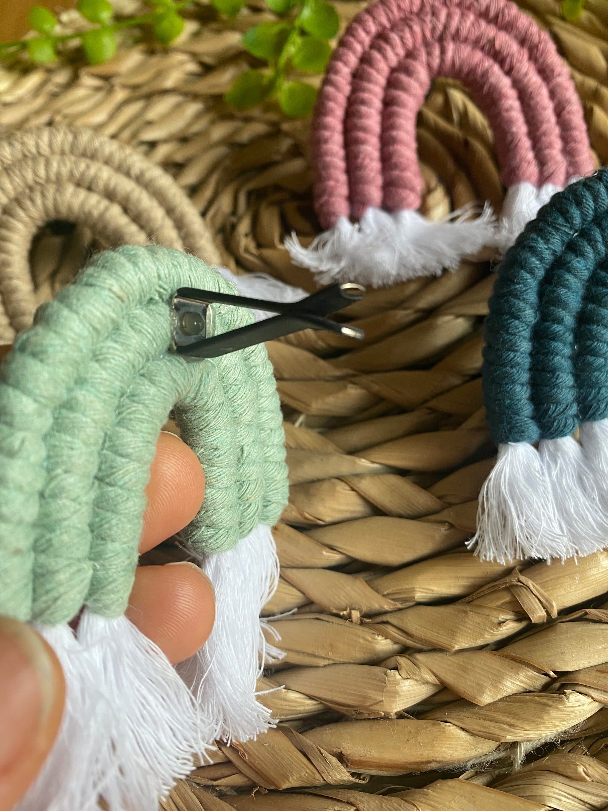 A hand touching a mini macrame rainbow in green, showing the black air vent clip on the back of the rainbow. There are three other mini rainbows on a wicker mat in the background.