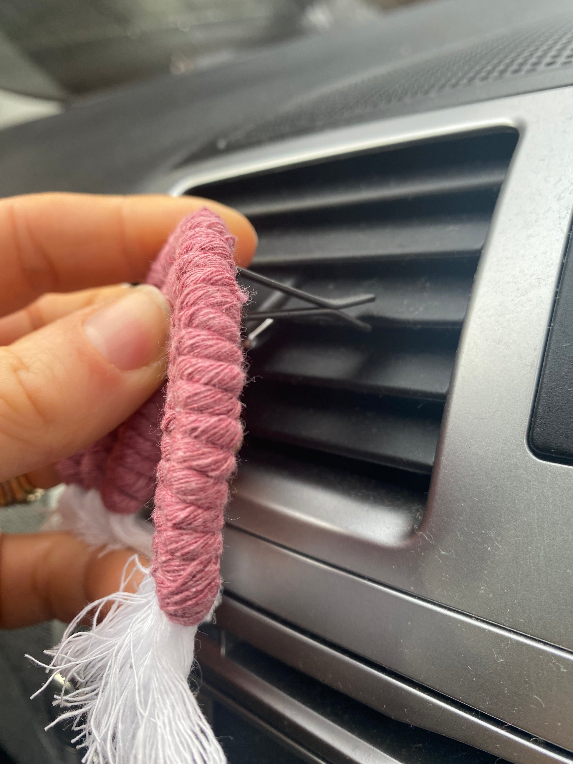 A hand attaching a mini macrame rainbow in pink, to the car air vent on a black car interior.