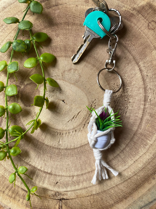 A miniature faux plant inside a mini macrame plant hanger, made with natural cotton, with a metal keyring clasp attached to a small set of keys, laid out on a wooden board next to some green plant vines.