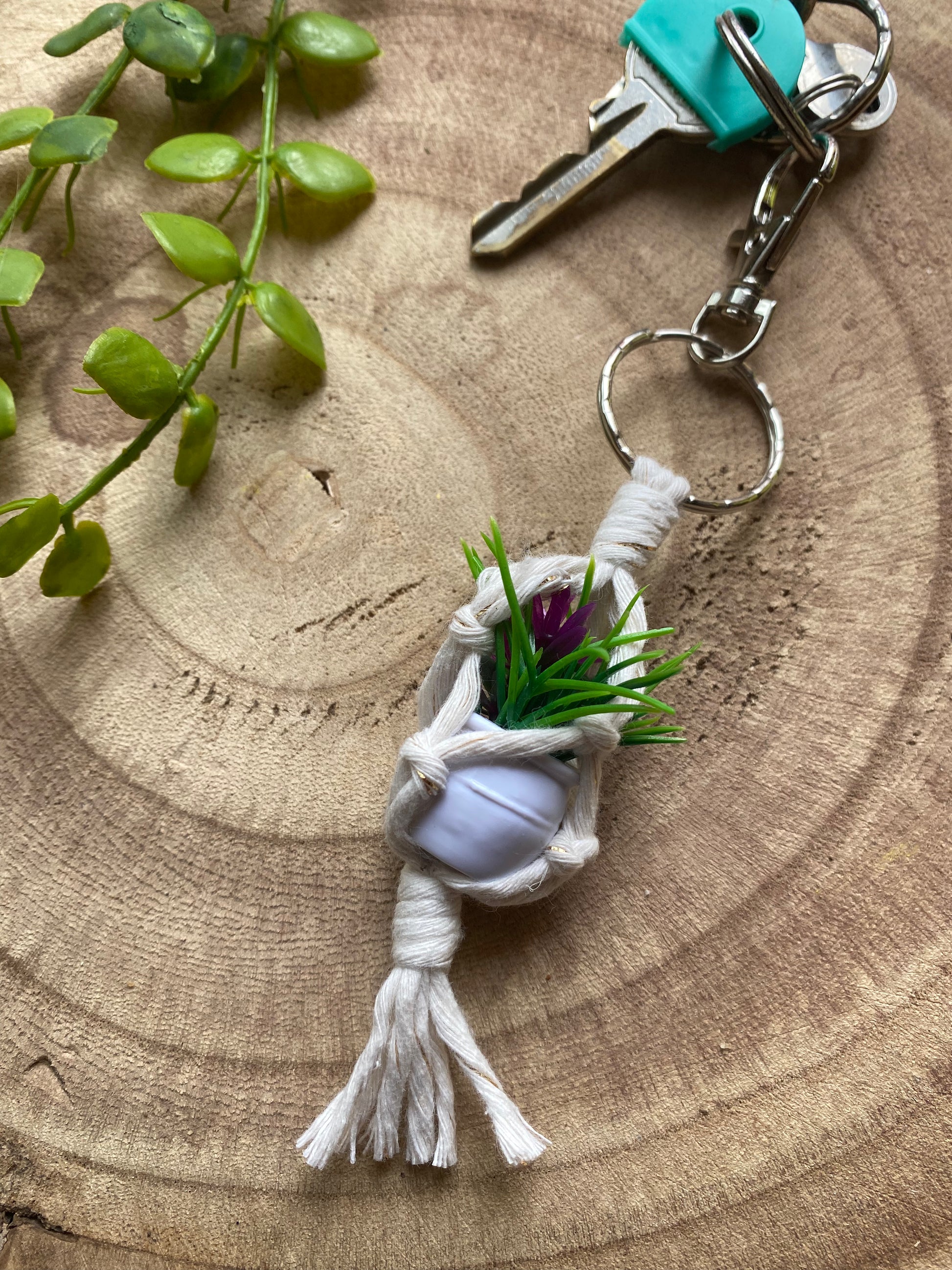 A miniature faux plant inside a mini macrame plant hanger, made with natural cotton, with a metal keyring clasp attached to a small set of keys, laid out on a wooden board next to some green plant vines.