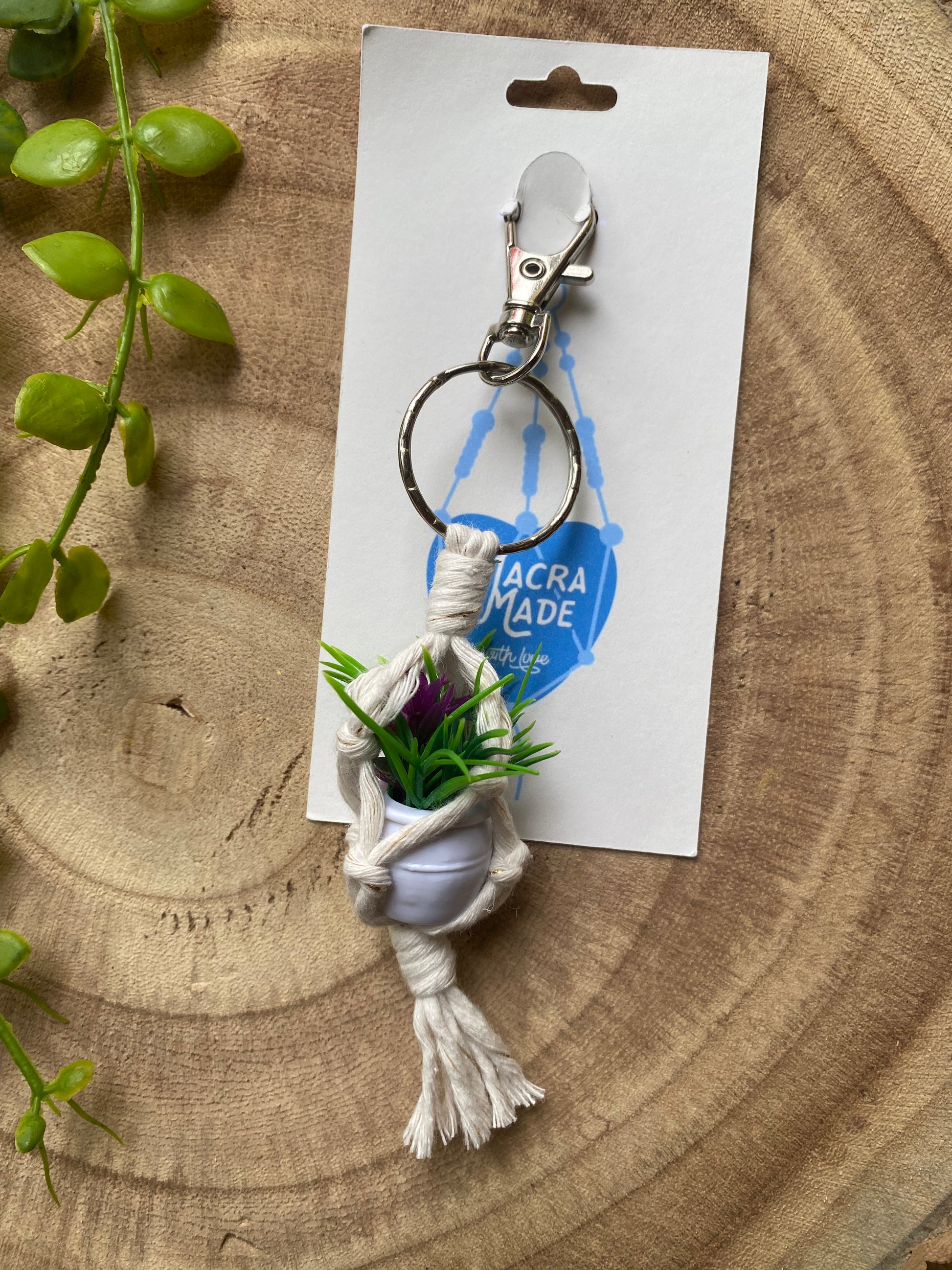 A miniature faux plant inside a mini macrame plant hanger, made with natural cotton, with a metal keyring clasp attached to a white cardboard tag, laid out on a wooden board next to some green plan vines.