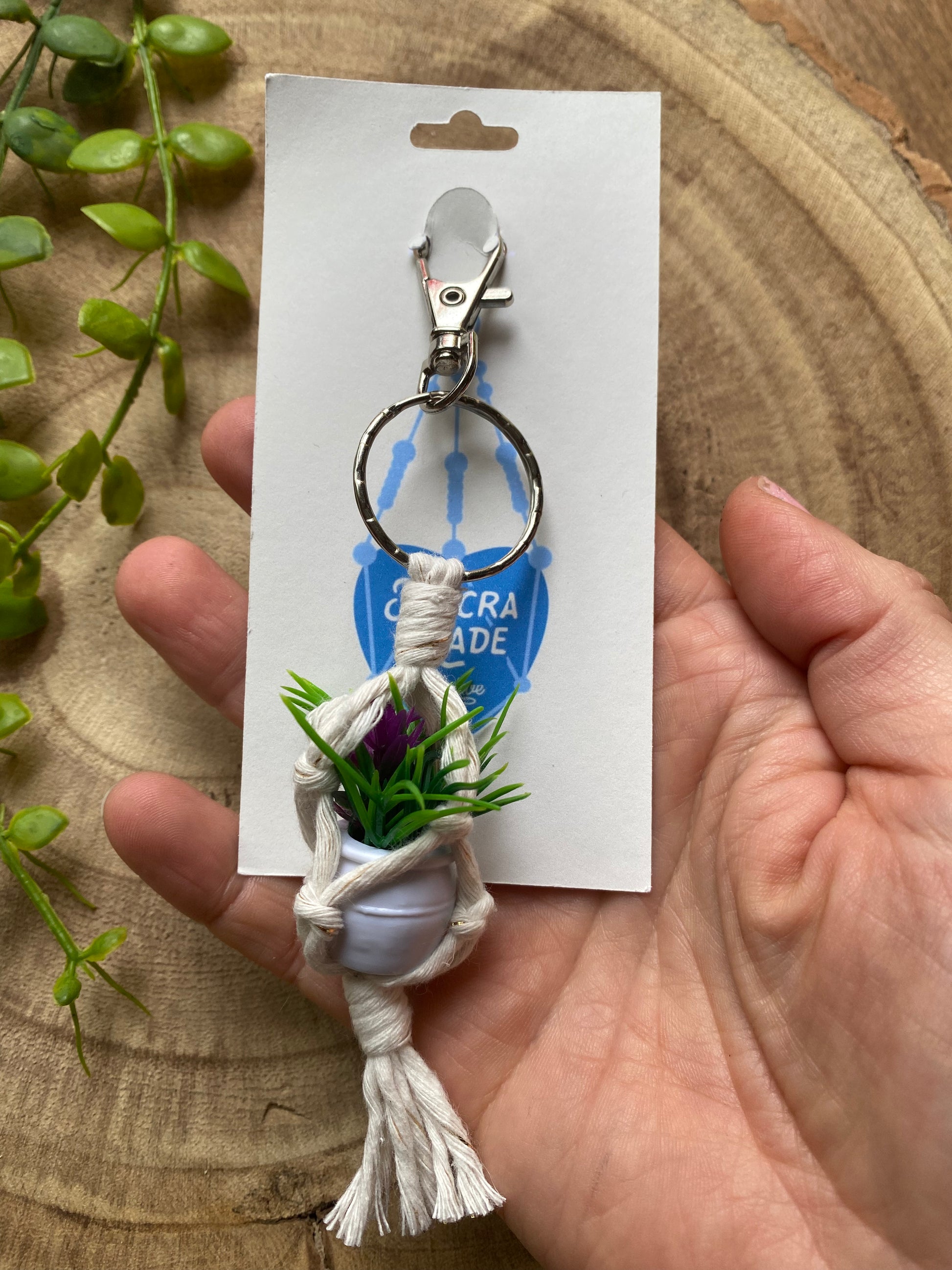 A hand holding a miniature faux plant hanger keyring attached to a white cardboard tag.
