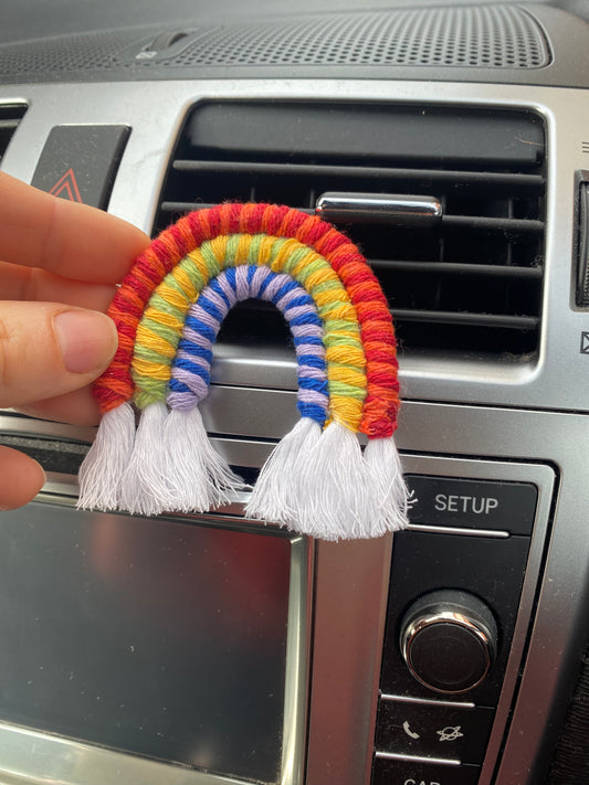A hand touching a mini macrame rainbow, attached to the car air vent on a black car interior.