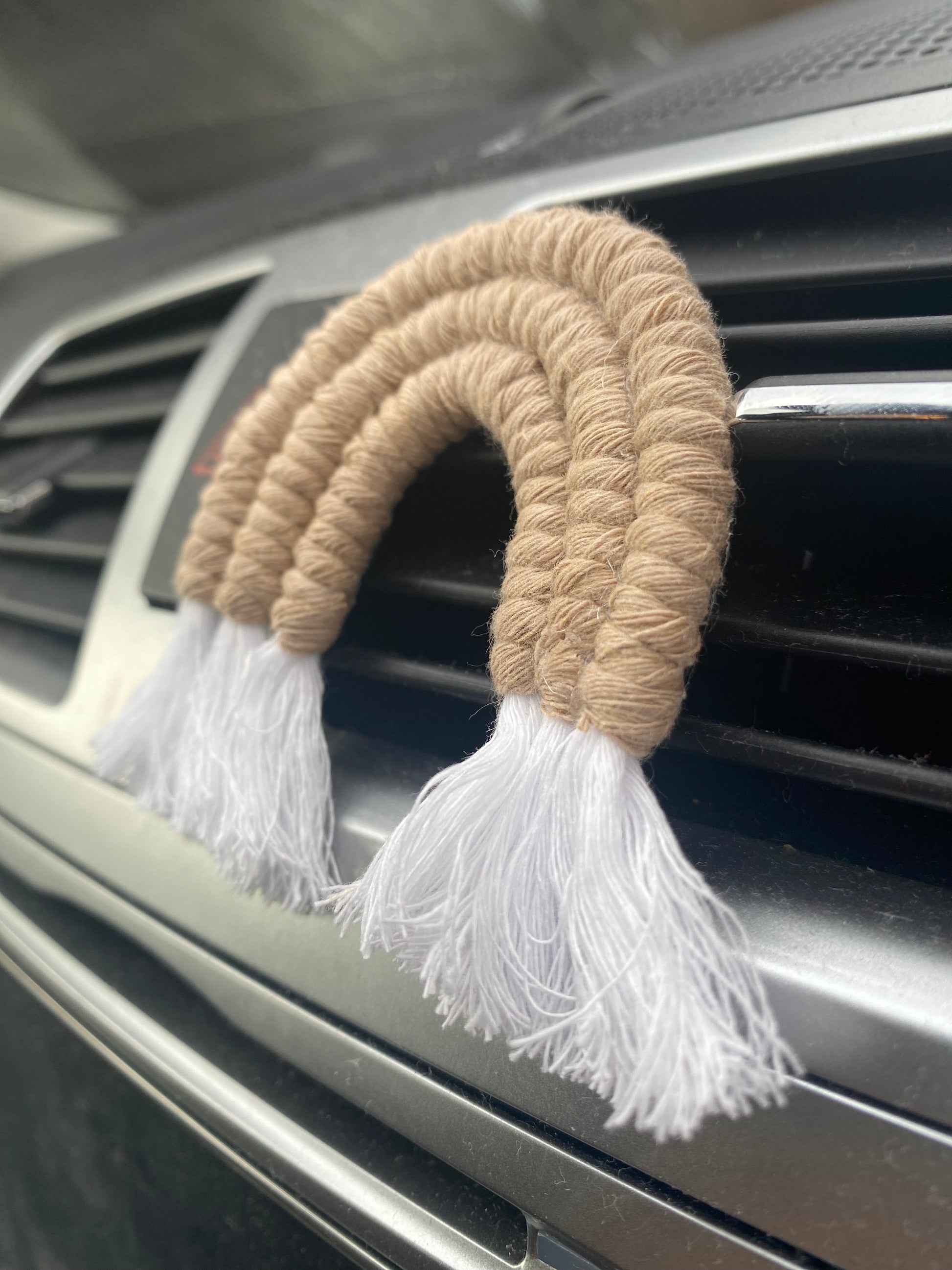 A mini macrame rainbow in beige, attached to the car air vent on a black car interior.