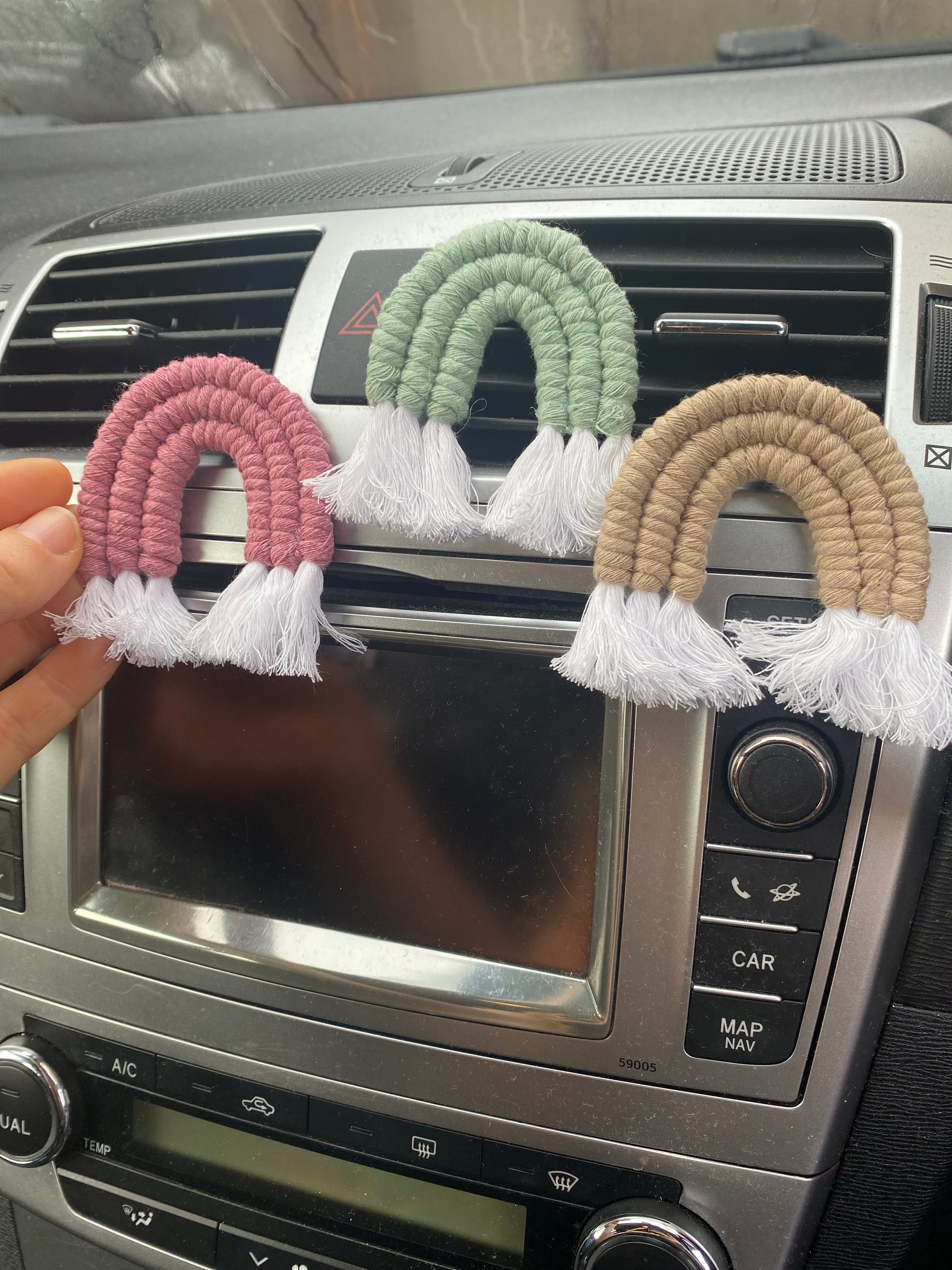 A hand touching a set of three mini macrame rainbows, attached to the car air vent on a black car interior. One rainbow is pink, one is green and one is beige.