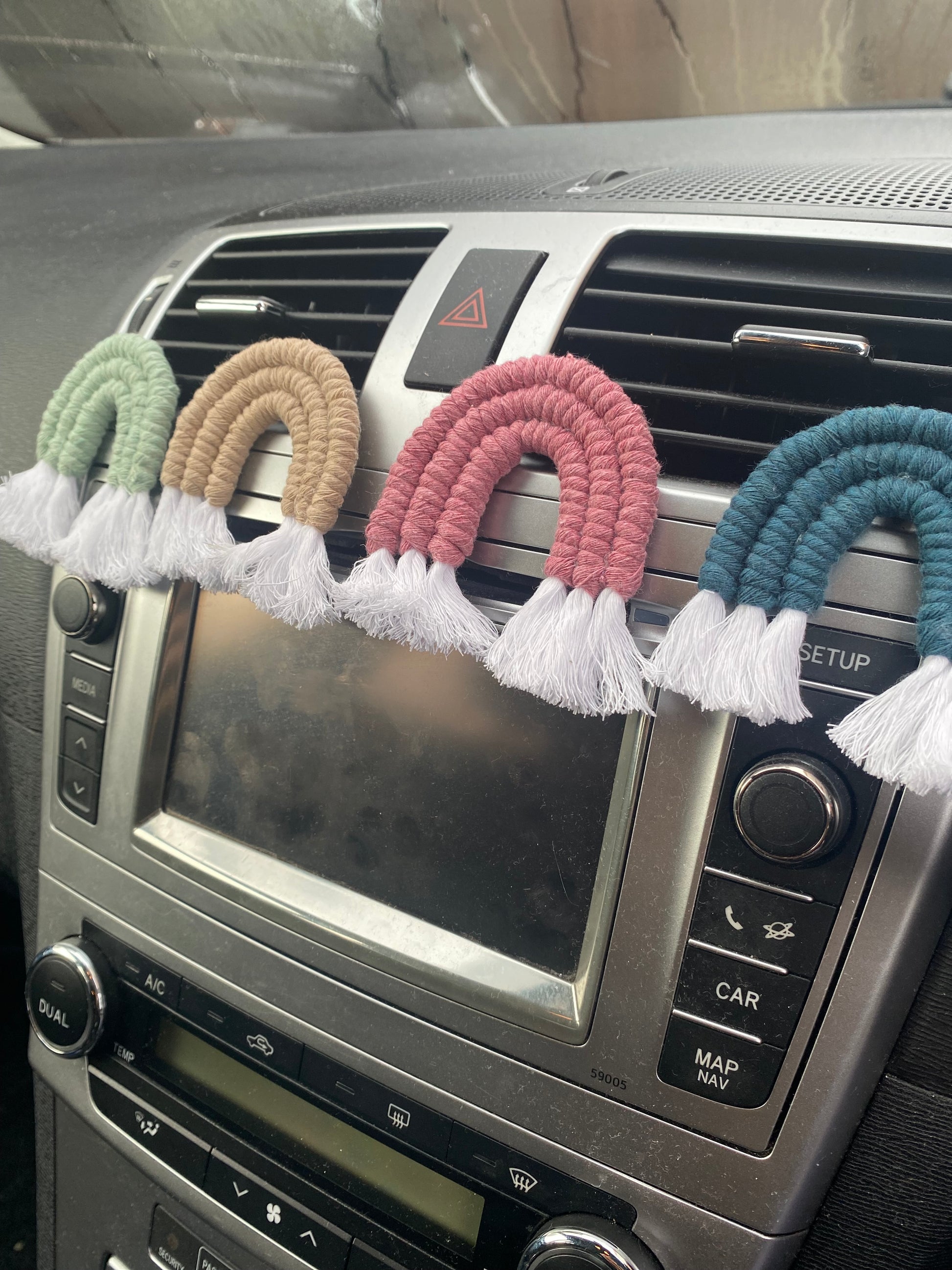 Four mini macrame rainbows in green, beige, pink and blue attached to the car air vent on a black car interior.