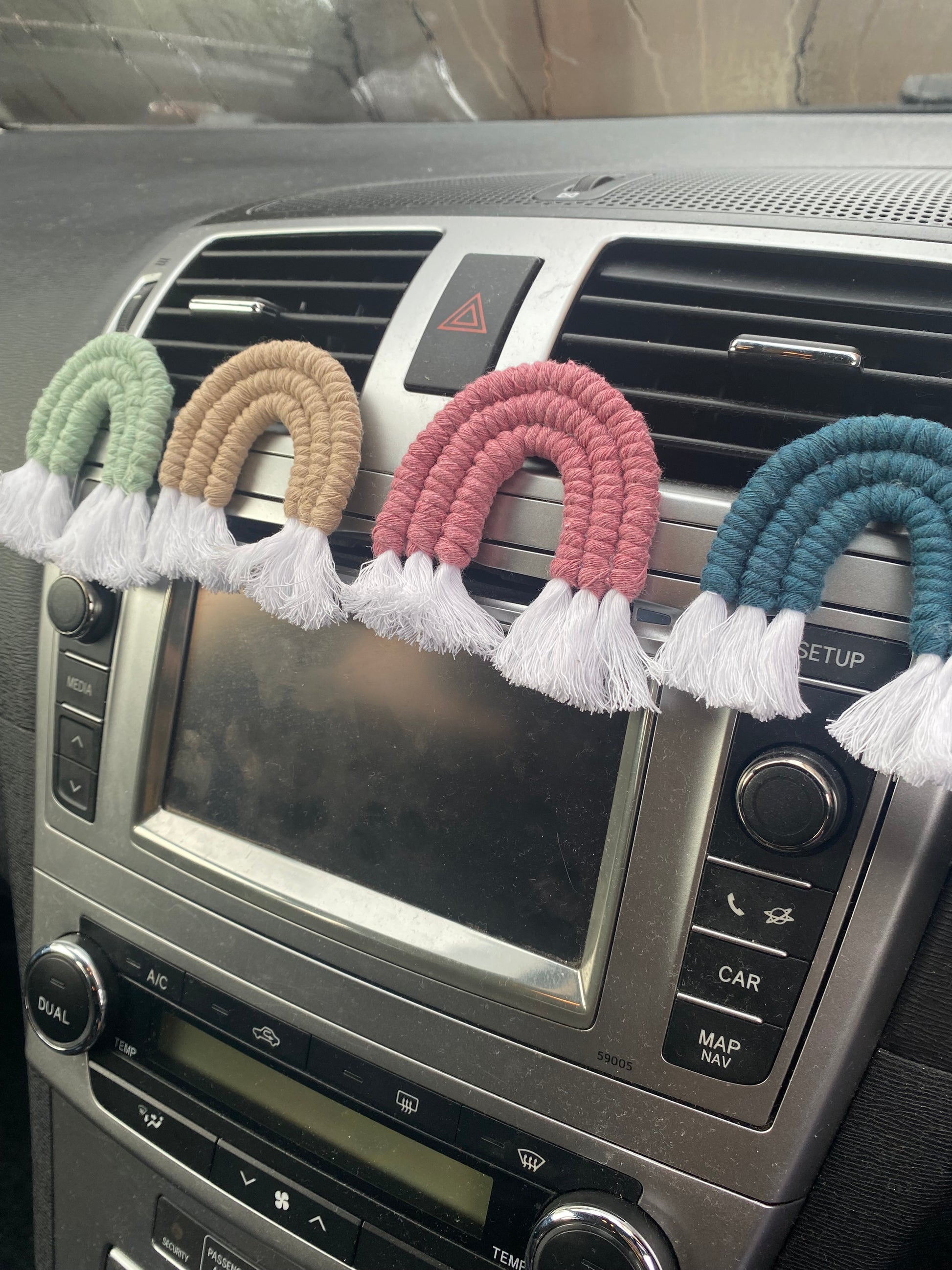 Four mini macrame rainbows in green, beige, pink and blue, attached to the car air vents on a black car interior.