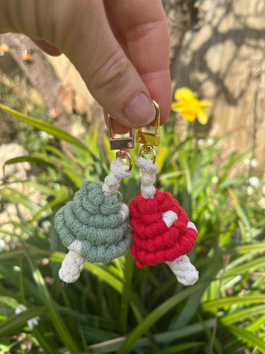 Two handmade macrame mushroom keyrings, made with eco friendly recycled cotton, one in green, one in red, each with shiny metal clasps. The keyring are being held outside in the sunshine, in front of green leafy plants.