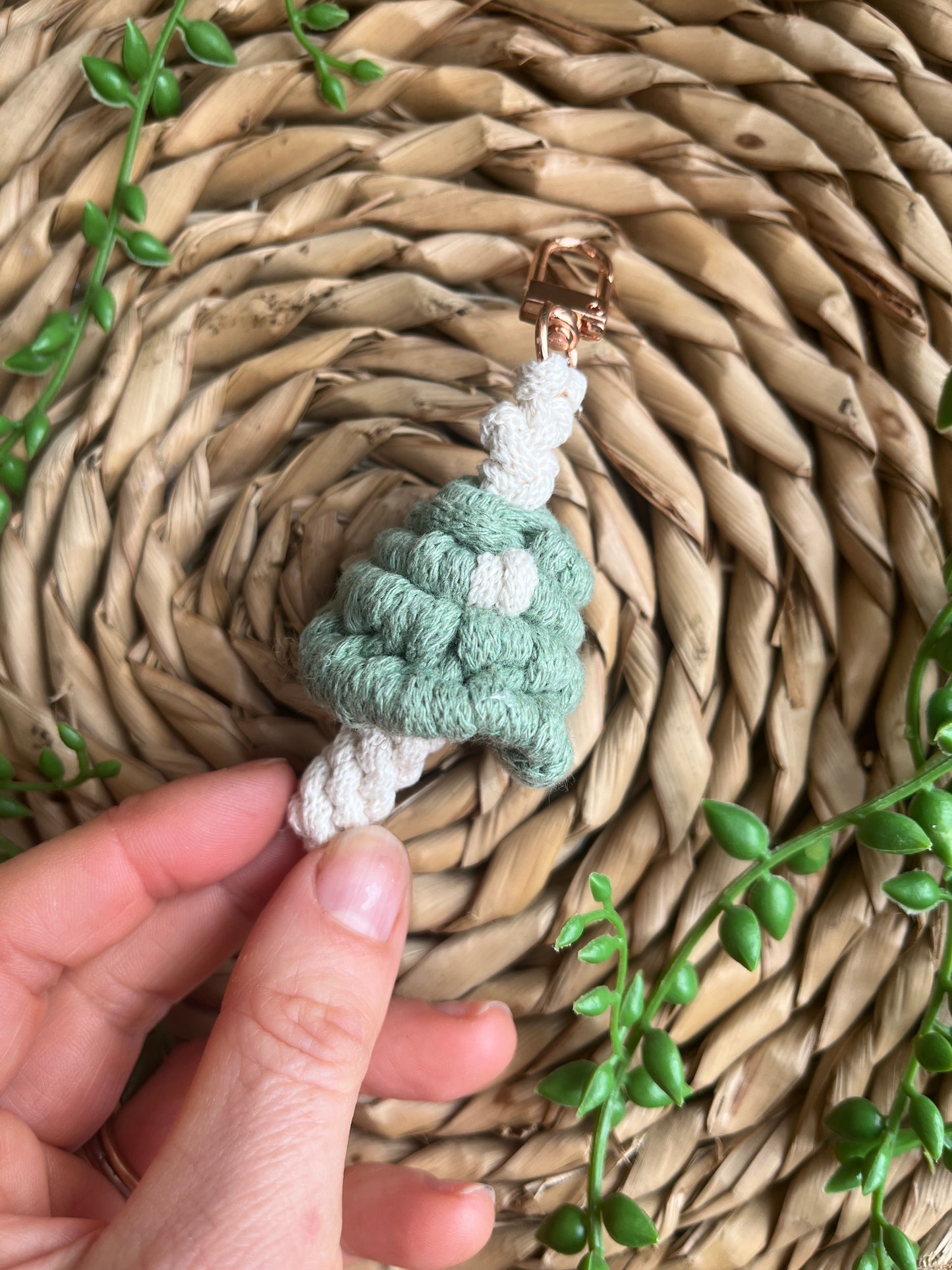 A hand holding a handmade macrame mushroom keyring, made with eco friendly recycled cotton in green, with a shiny metal clasp. The keyring is displayed on a wicker mat, with some long green plant vines.