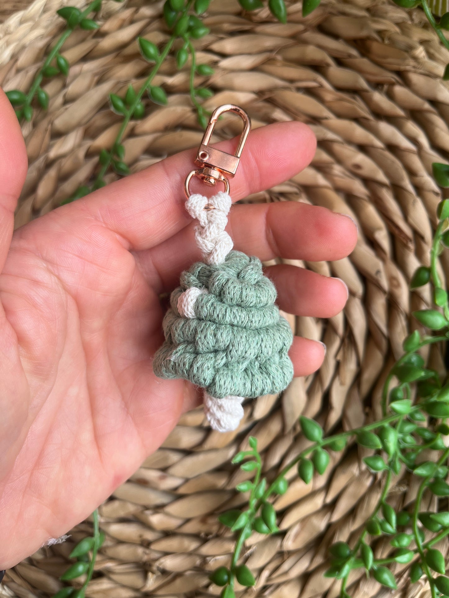 A hand holding a handmade macrame mushroom keyring, made with eco friendly recycled cotton in green, with a shiny metal clasp. The keyring is displayed on a wicker mat, with some long green plant vines.