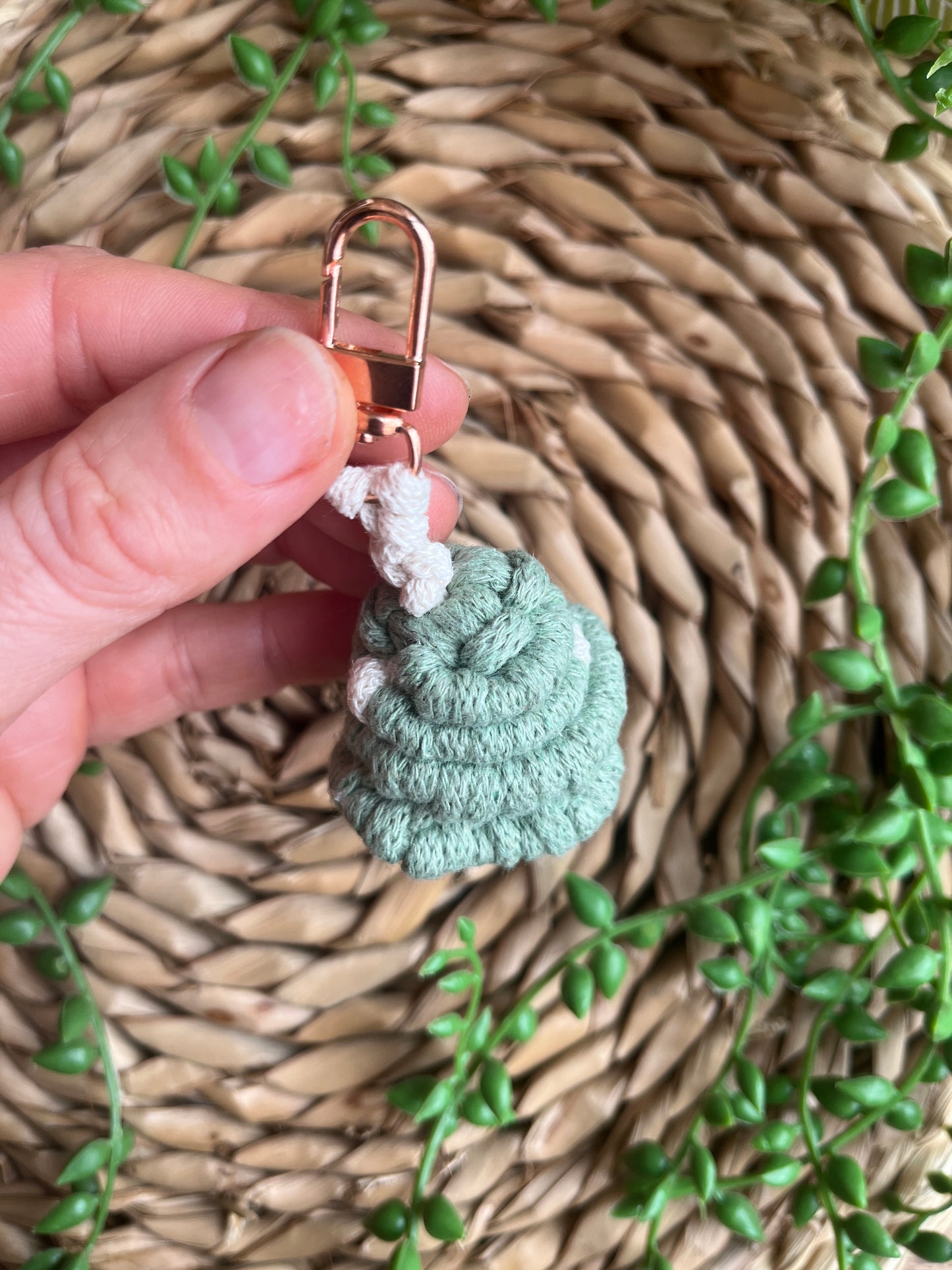 A hand holding a handmade macrame mushroom keyring, made with eco friendly recycled cotton in green, with a shiny metal clasp. The keyring is displayed on a wicker mat, with some long green plant vines.