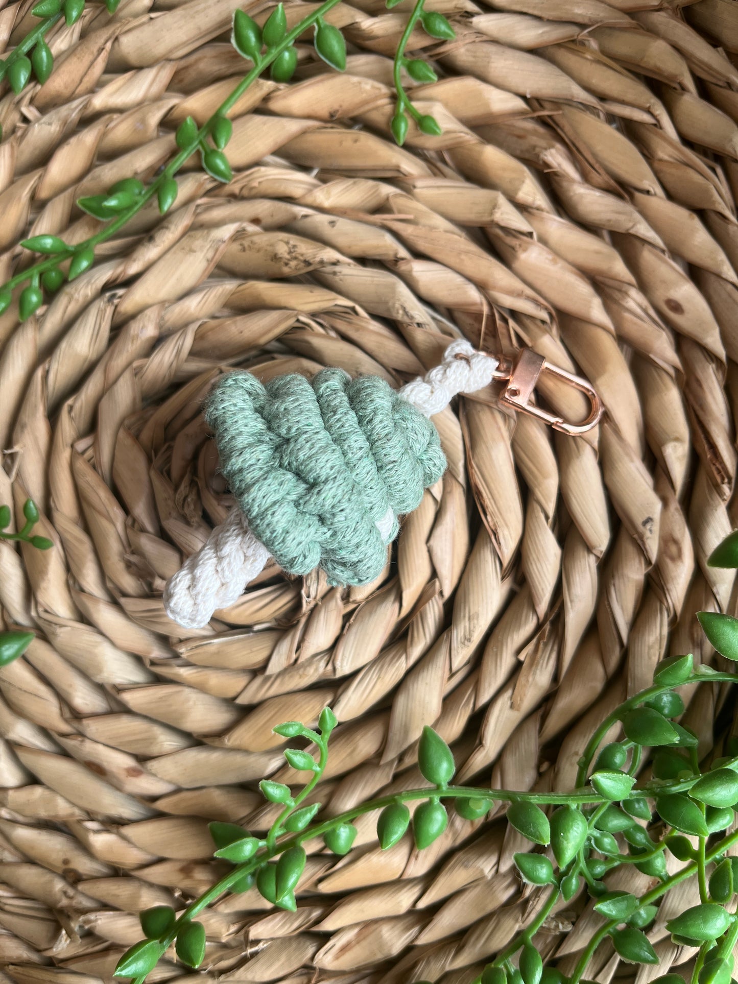 A handmade macrame mushroom keyring, made with eco friendly recycled cotton in green, with a shiny metal clasp. The keyring is displayed on a wicker mat, with some long green plant vines.