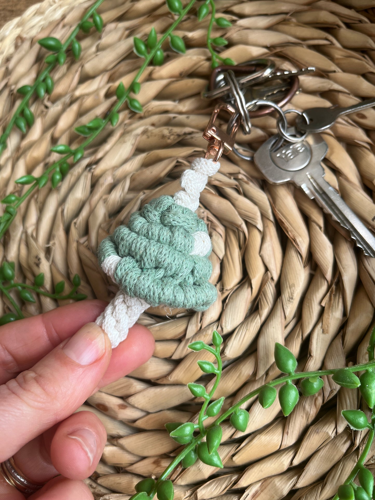 A hand holding a handmade macrame mushroom keyring, made with eco friendly recycled cotton in green, with a shiny metal clasp. The keyring is displayed on a wicker mat, with some long green plant vines attached to a small set of silver keys.