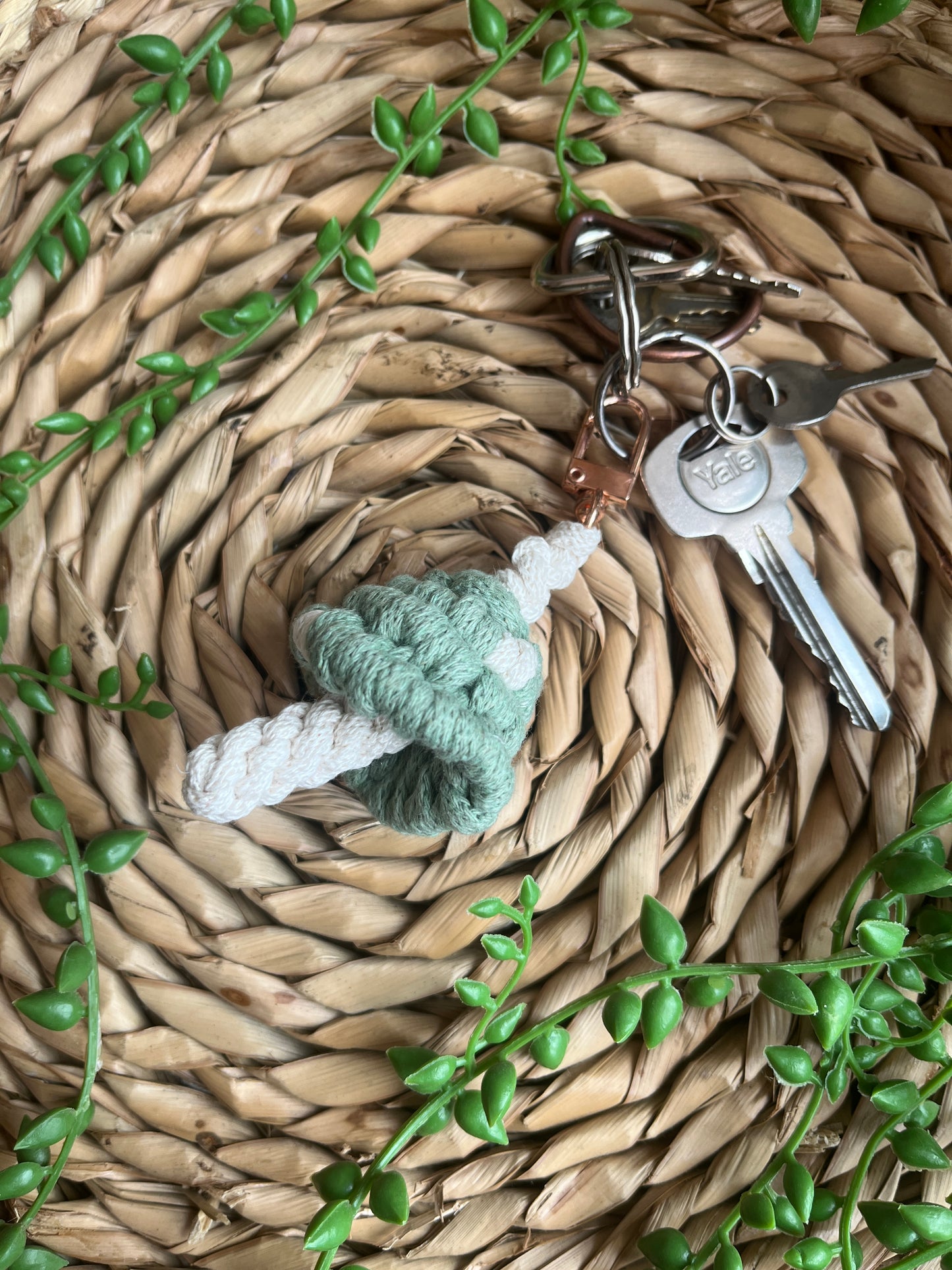 A handmade macrame mushroom keyring, made with eco friendly recycled cotton in green, with a shiny metal clasp. The keyring is displayed on a wicker mat, with some long green plant vines attached to a small set of silver keys.