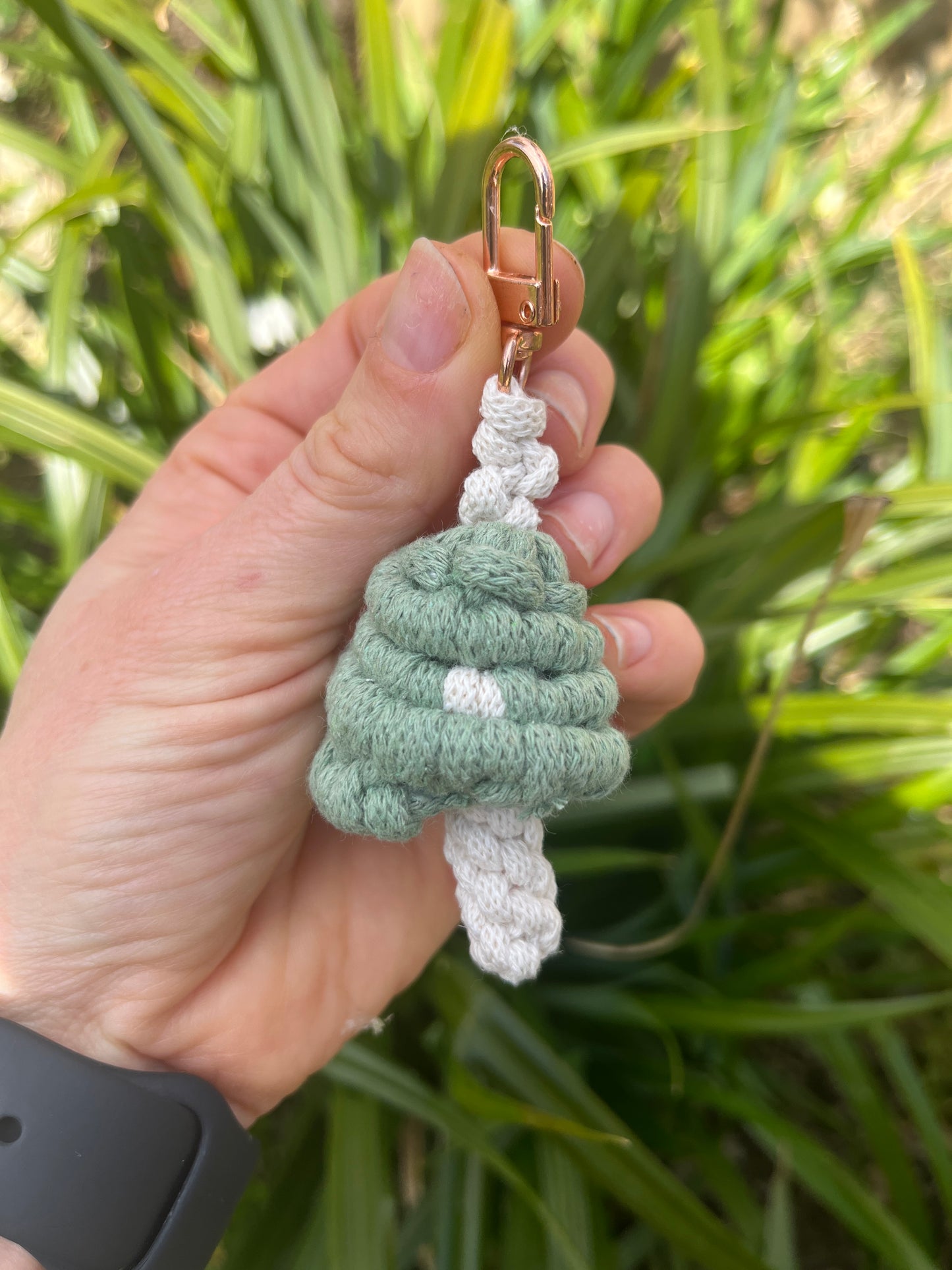 A hand holding a handmade macrame mushroom keyring, made with eco friendly recycled cotton in green, with a shiny metal clasp. The keyring is being held outside in front of some green leafy plants.