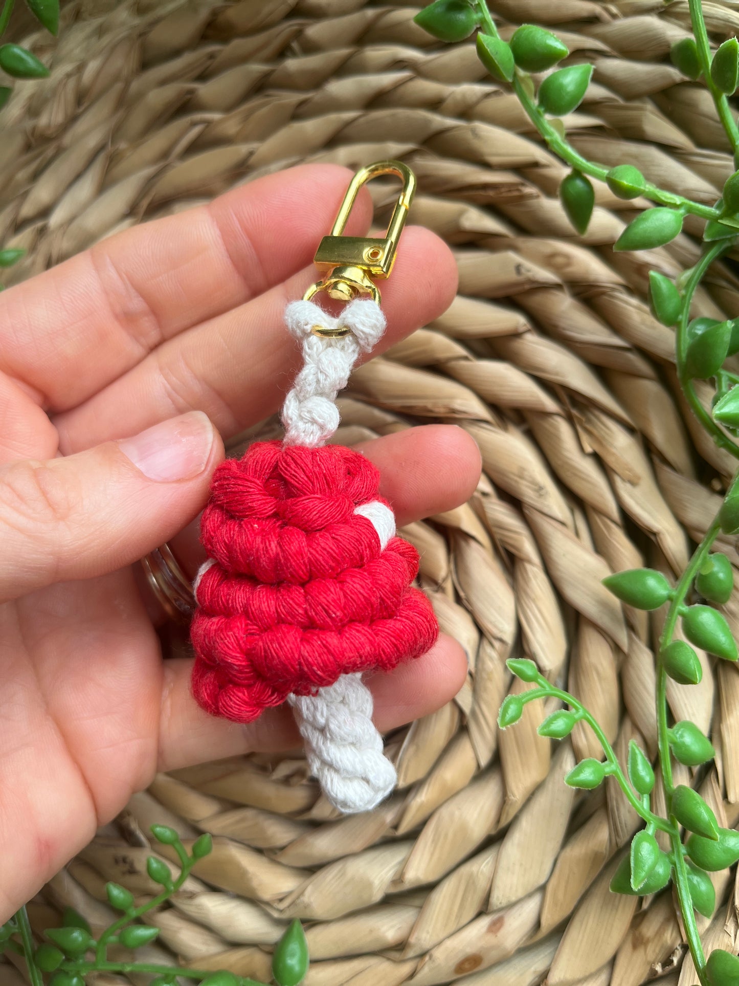 A hand is holding a handmade macrame mushroom keyring, made with eco friendly recycled cotton in red, with a shiny metal clasp. The keyring is displayed on a wicker mat, with some long green plant vines.