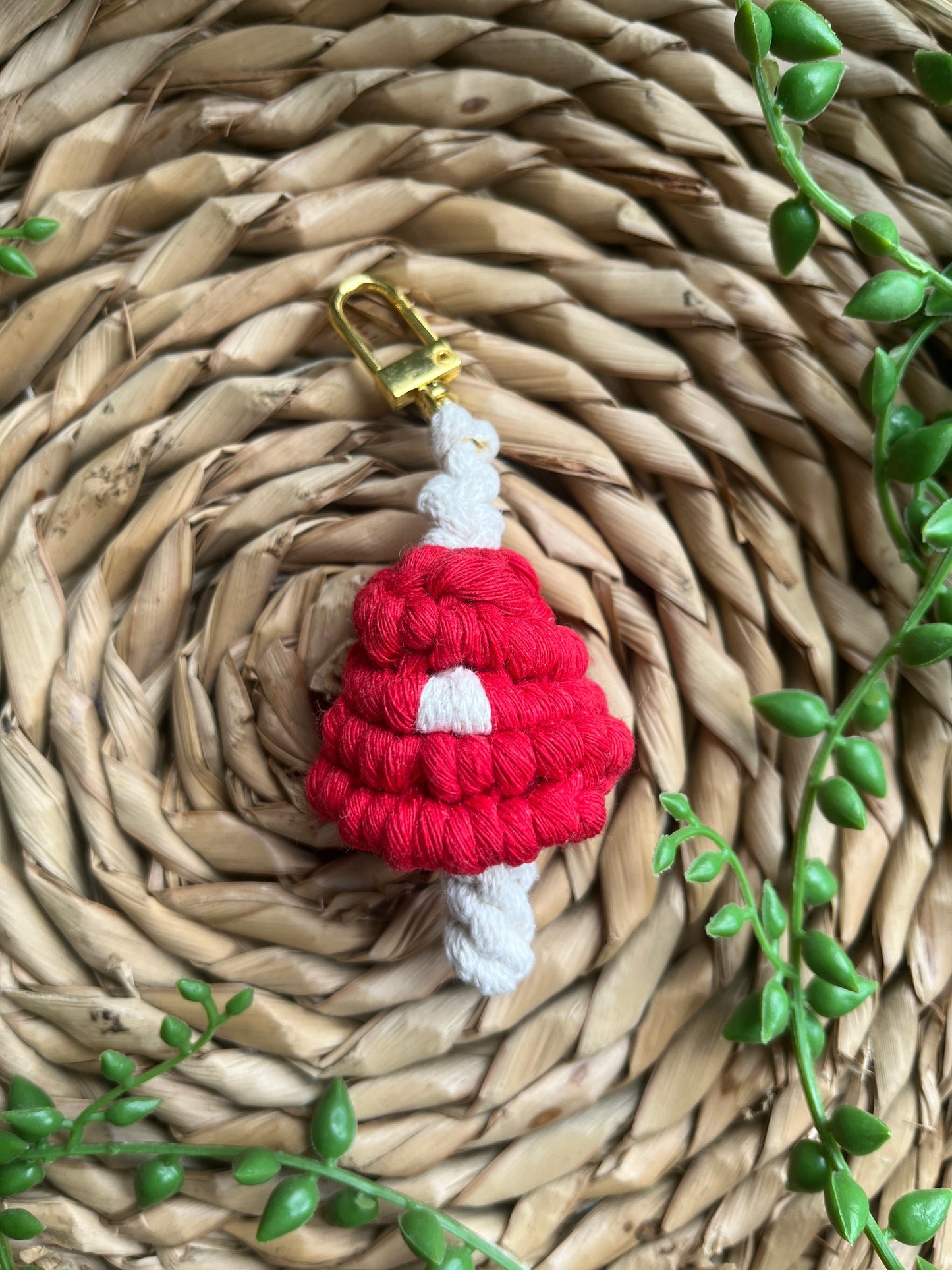 A handmade macrame mushroom keyring, made with eco friendly recycled cotton in red, with a shiny metal clasp. The keyring is displayed on a wicker mat, with some long green plant vines.