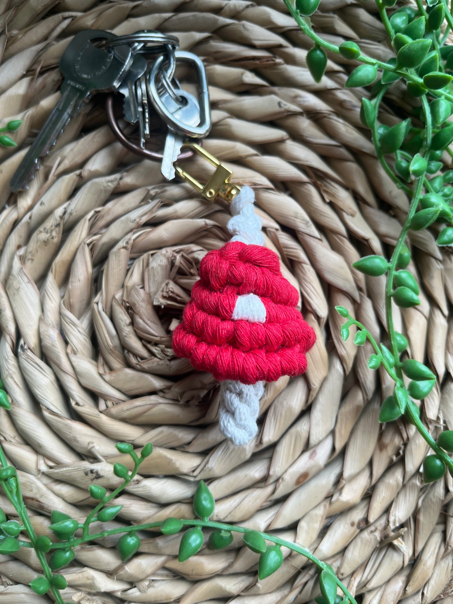 A handmade macrame mushroom keyring, made with eco friendly recycled cotton in red, with a shiny metal clasp. The keyring is displayed on a wicker mat, with some long green plant vines, attached to a small set of silver keys.