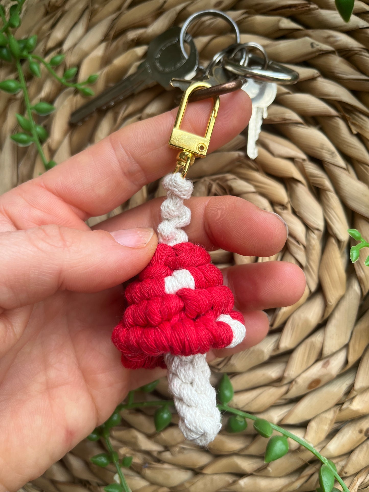 A hand is holding a handmade macrame mushroom keyring, made with eco friendly recycled cotton in red, with a shiny metal clasp. The keyring is displayed on a wicker mat, with some long green plant vines attached to a small set of silver keys.