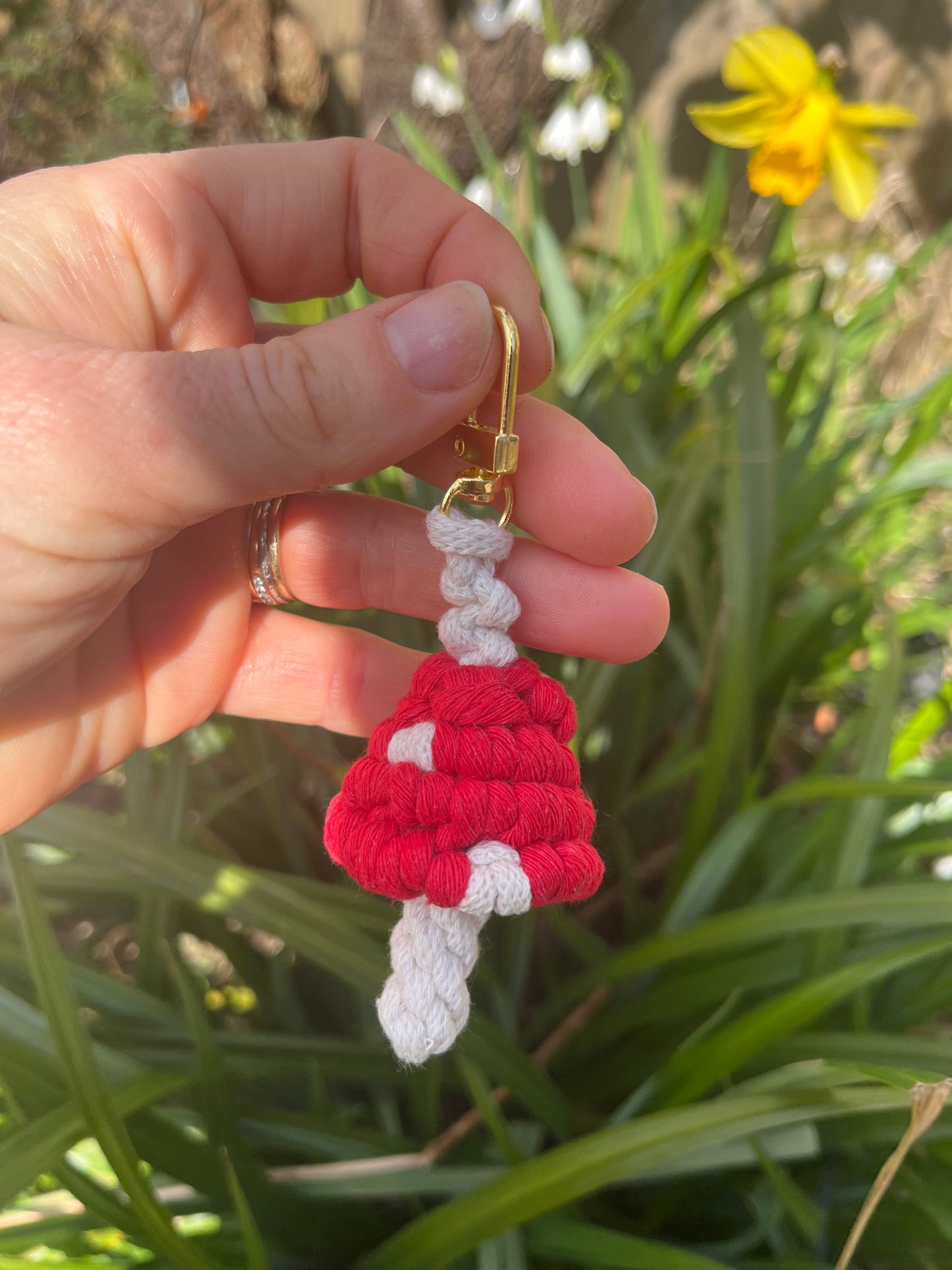 A had holding a handmade macrame mushroom keyring, made with eco friendly recycled cotton in red, with a shiny metal clasp. The keyring is being held outside in front of some green leafy plants.