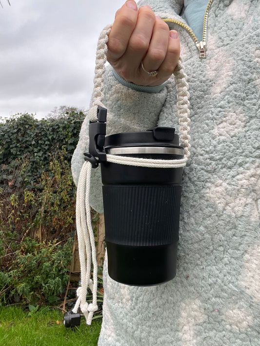 Adjustable coffee cup holder strap made with eco friendly natural cotton, demonstrated with a black travel mug, being held outside with leafy green plants in the background.