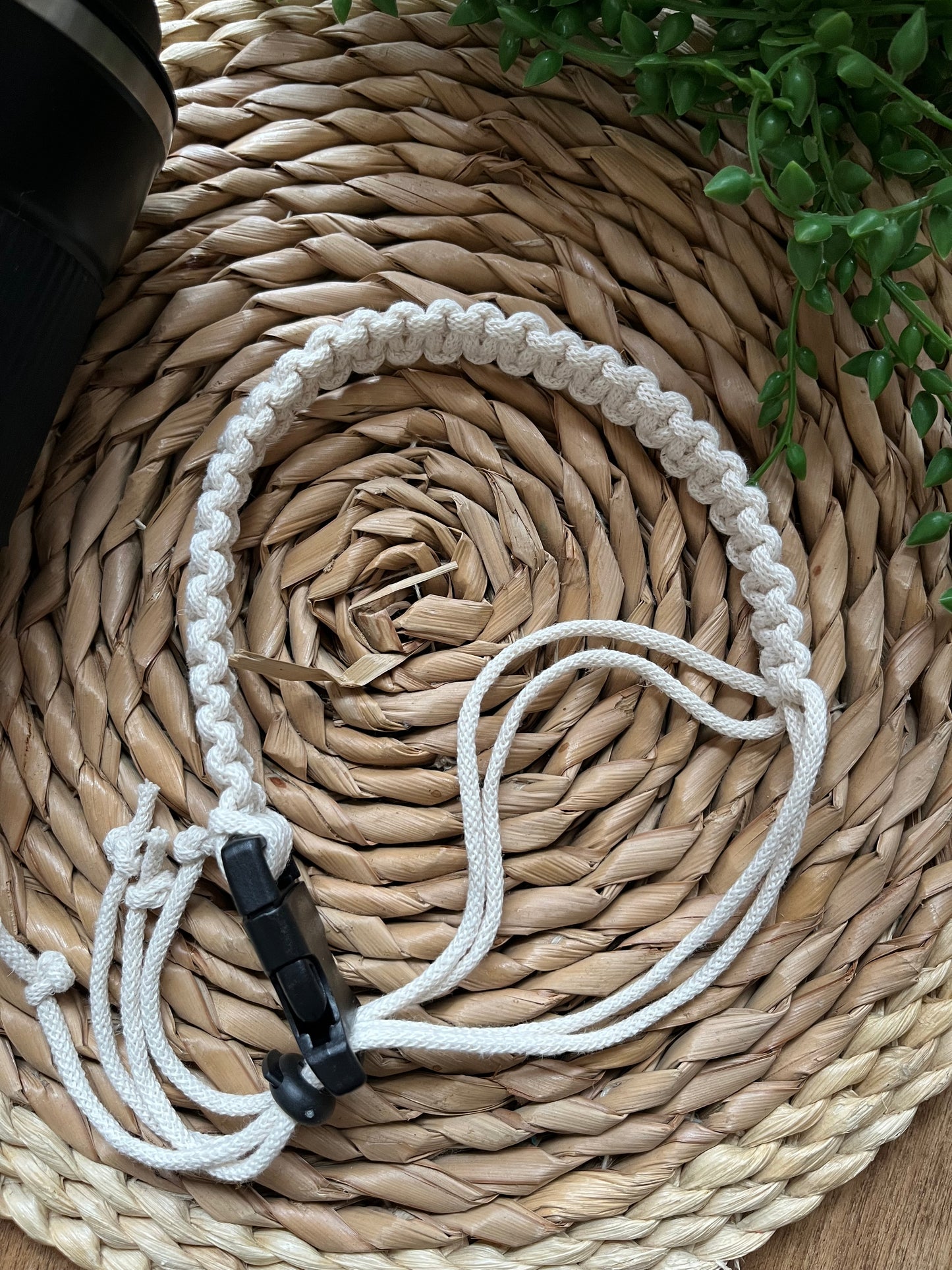 A handmade macrame coffee cup holder strap lays on a wicker mat, with some green plant vines and a reusable travel cup. The strap features neat knots, made with natural recycled cotton, and has a black buckle to fasten and a black toggle to adjust.