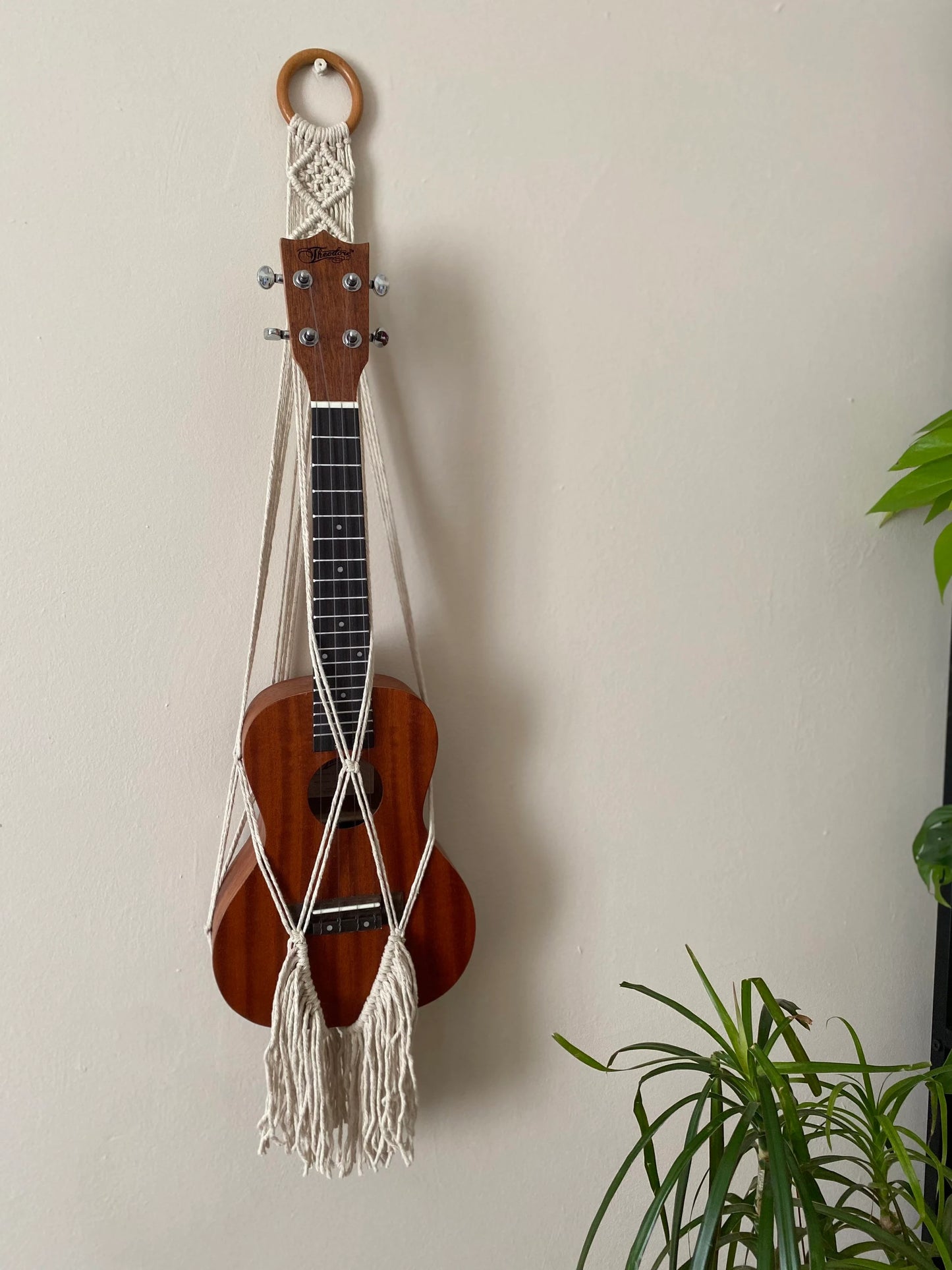 A dark brown ukulele sits inside of a handmade macrame wall hanging ukulele hanger, which is made with natural recycled cotton with a long boho fringe, and a repurposed wooden hanging ring. Green leafy plants are visible to the right of the image.