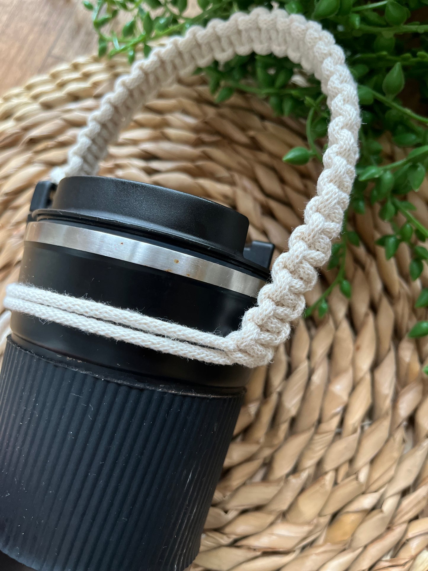 A black reusable coffee cup is laying on a wicker mat, next to some green plant vines, with a short natural handmade coffee cup holder strap attached to it.