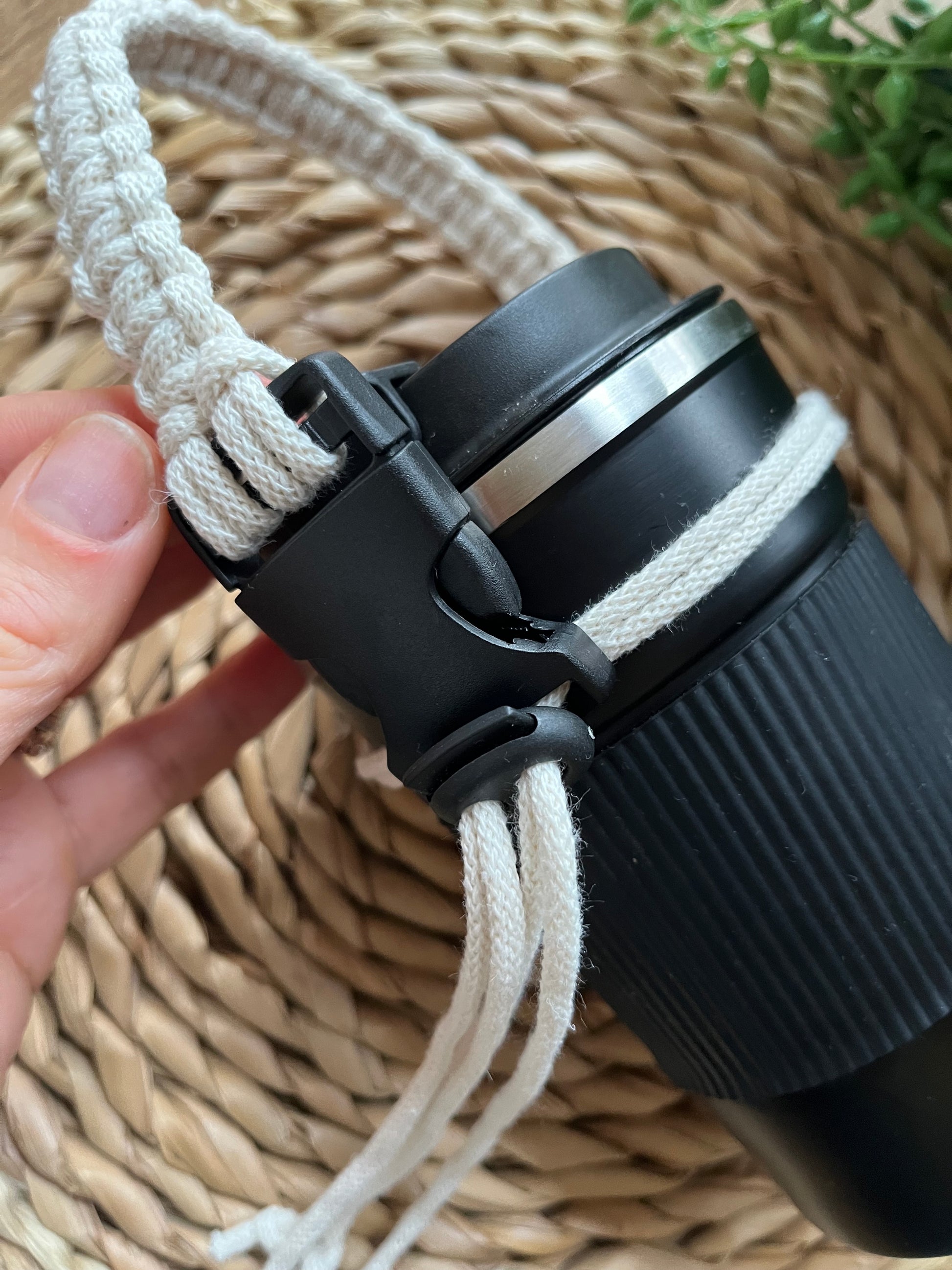 A hand holds a macrame coffee cup holder strap, which is attached to a black reusable coffee cup. The strap is made with natural recycled cotton and has a black clasp and toggle.