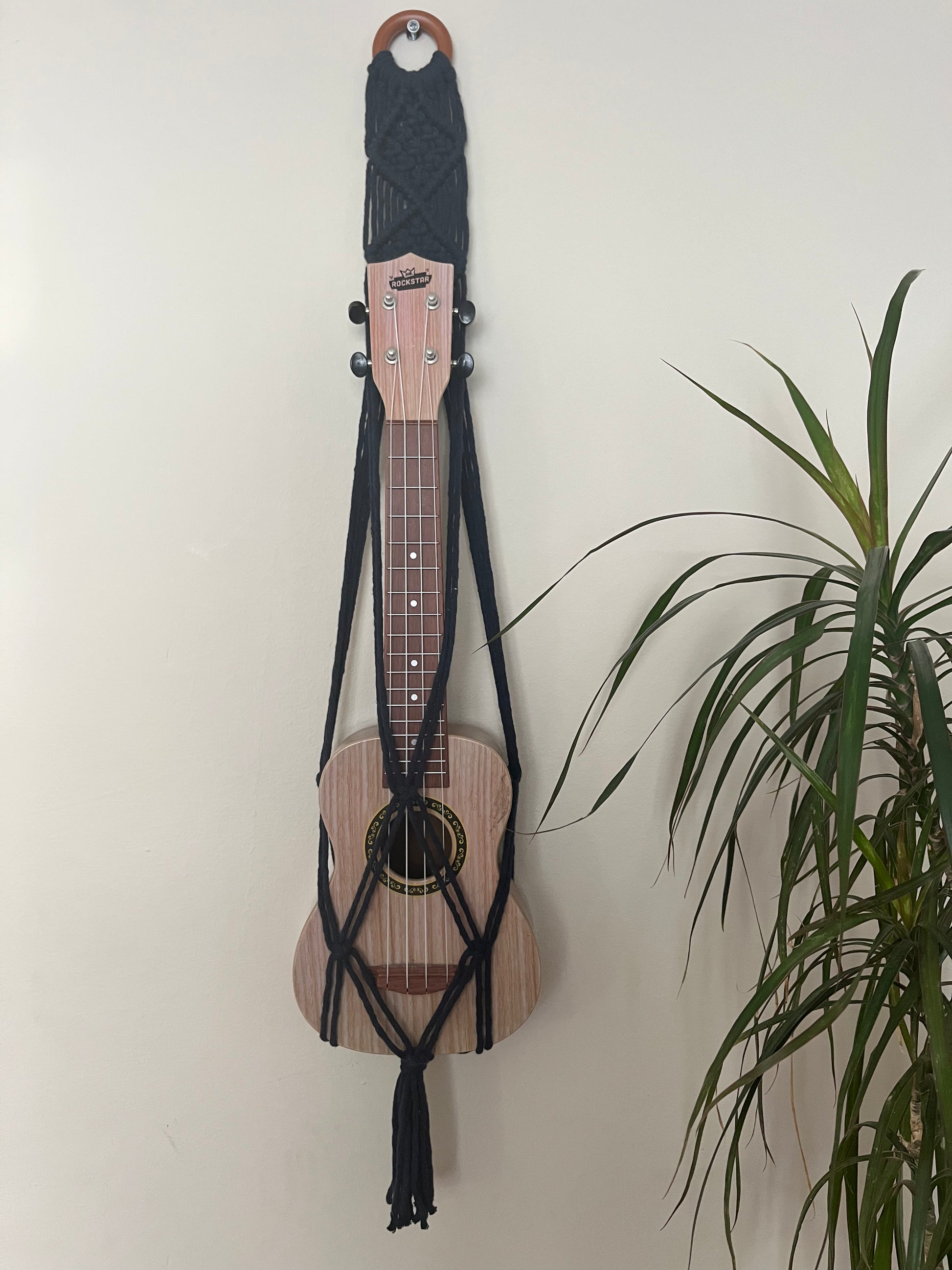 A light brown ukulele sits inside of a handmade macrame wall hanging ukulele hanger, which is made with navy blue recycled cotton, and a repurposed wooden hanging ring. A large green leafy plant is to the right of the image.