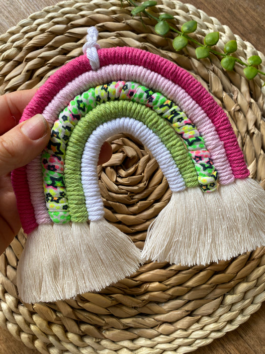 A hand holding a macrame rainbow with 5 arches of pink, green and neon coloured yarns and natural combed out tassels at the bottom.