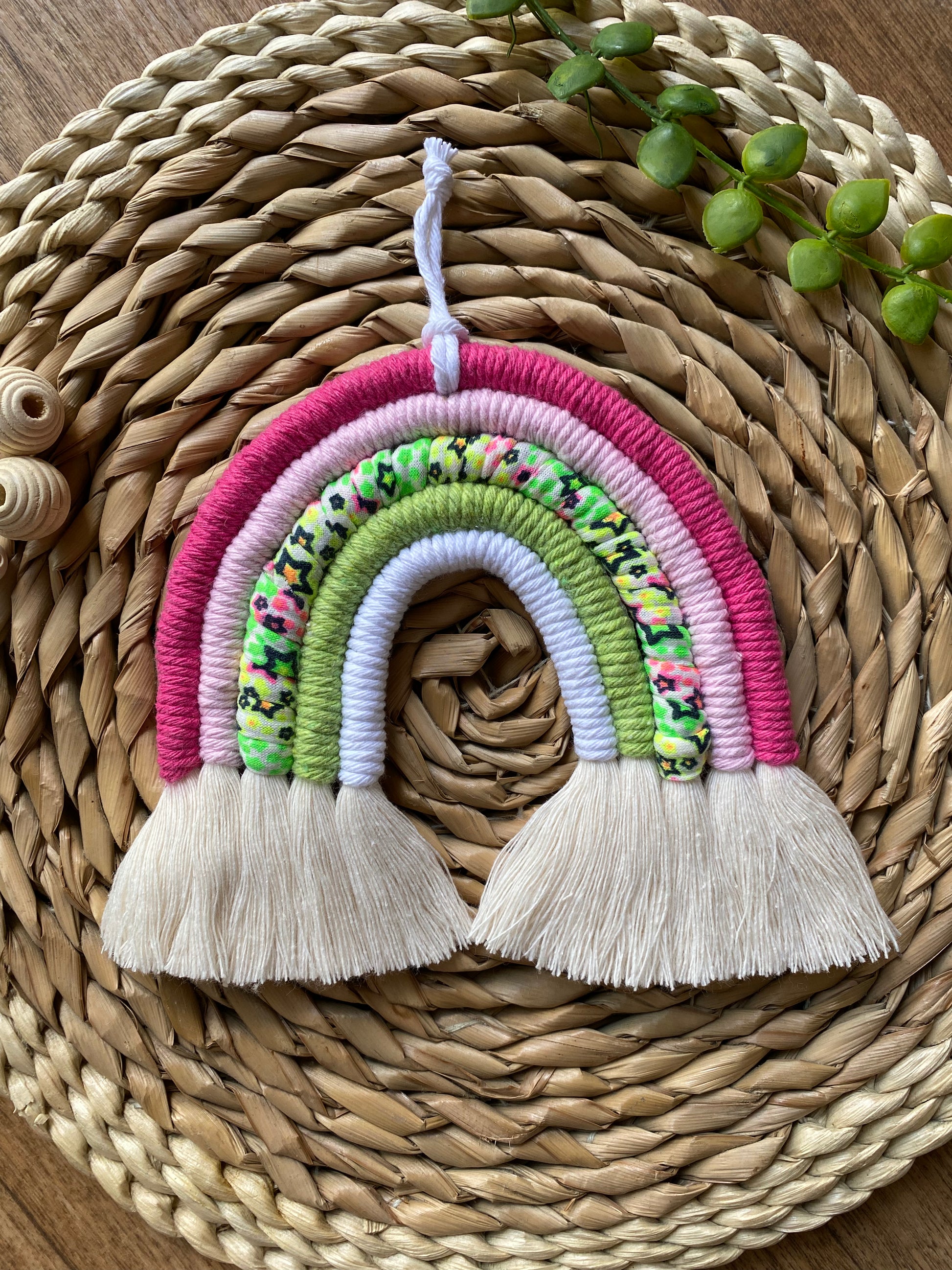 A macrame rainbow with 5 arches of pink, green and neon coloured yarns and natural combed out tassels at the bottom on a wicker mat with green plant vines and wooden beads for decoration.