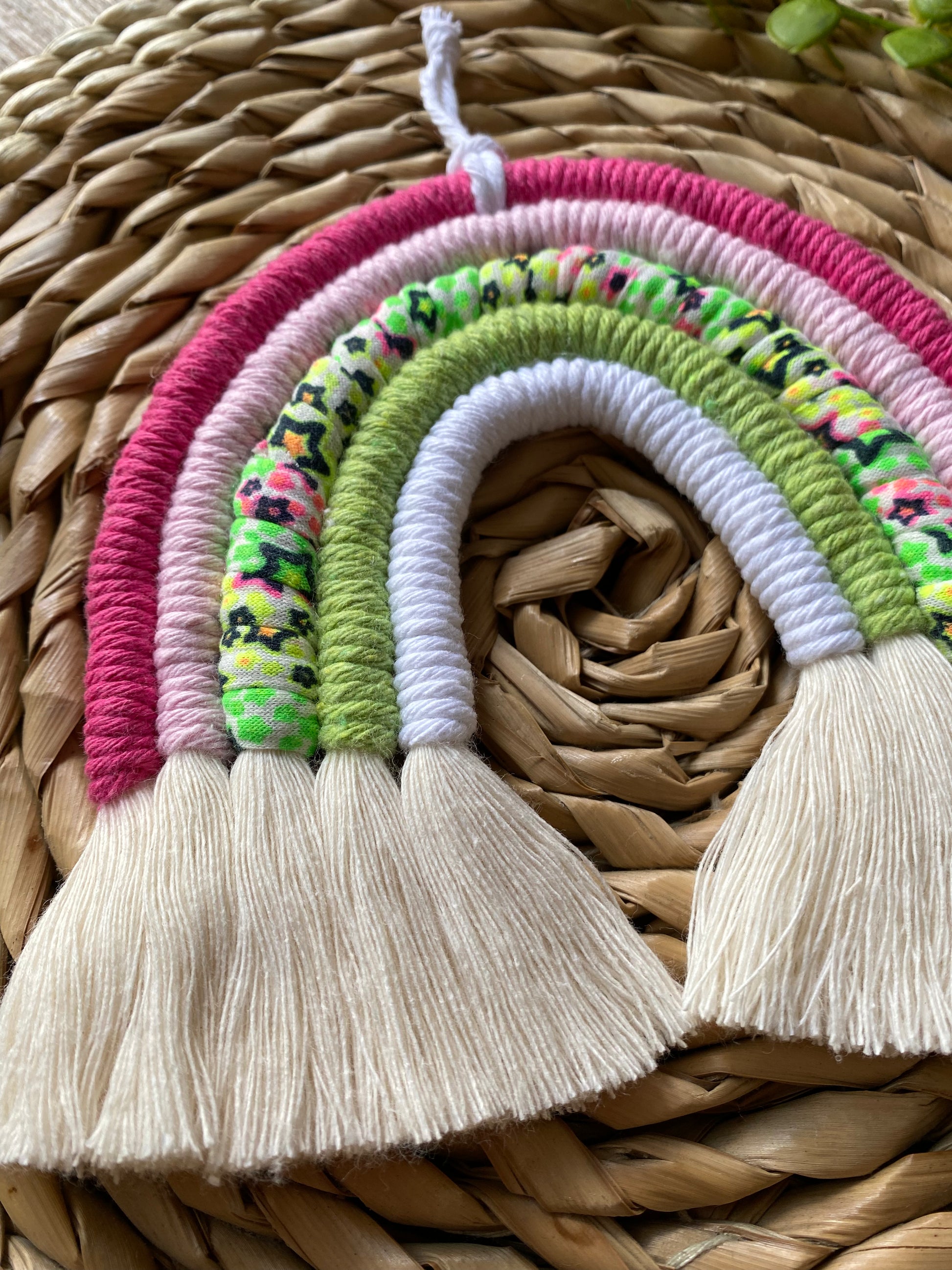 A close up of a macrame rainbow with 5 arches of pink, green and neon coloured yarns and natural combed out tassels at the bottom on a wicker mat.
