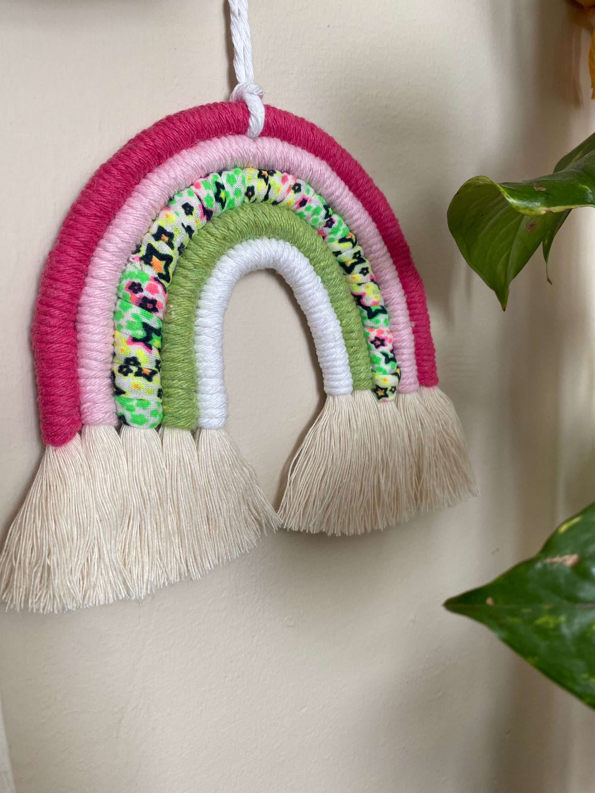 A macrame rainbow with 5 arches of pink, green and neon coloured yarns and natural combed out tassels at the bottom hanging against a white wall.