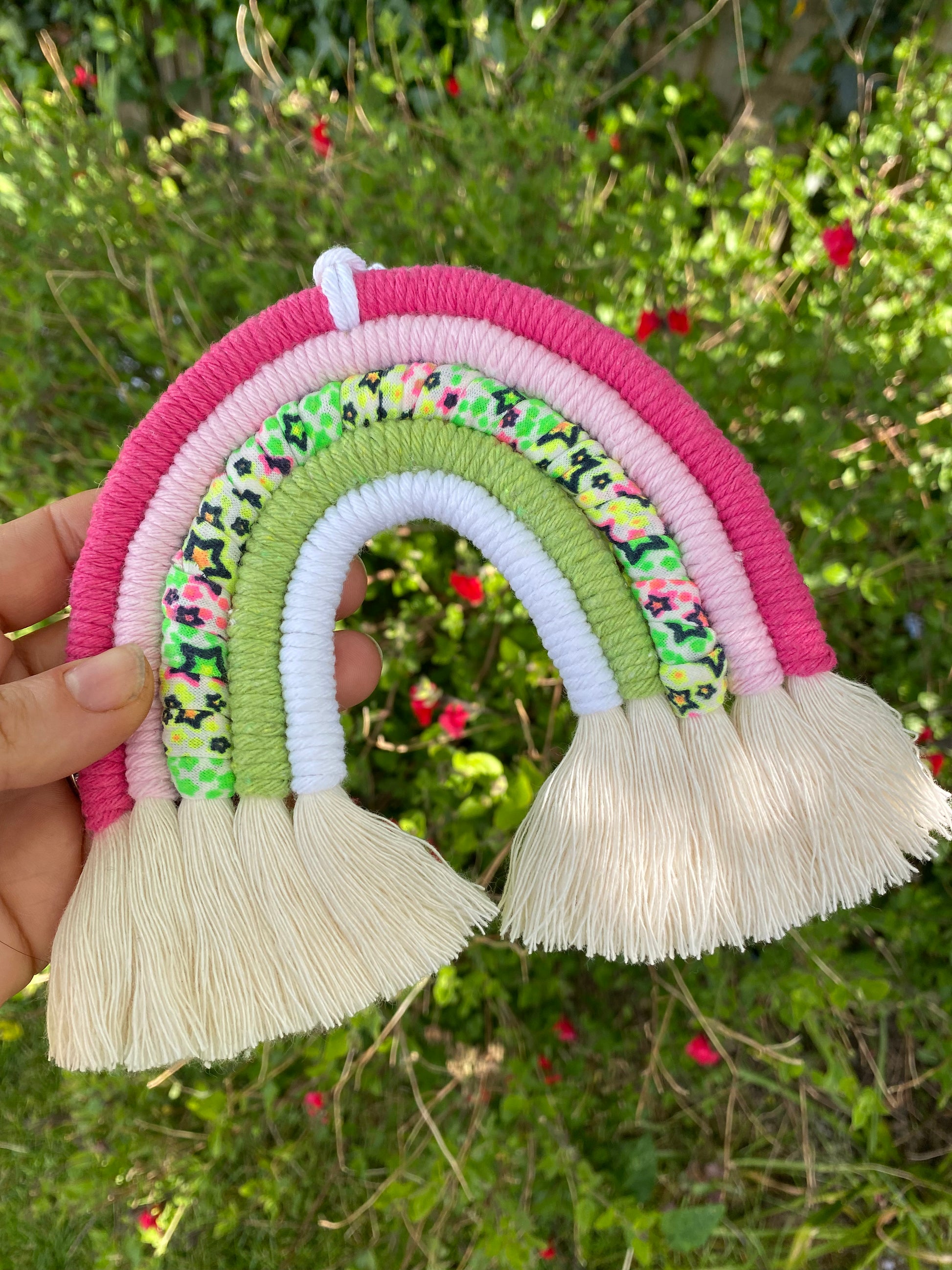 A hand holding a macrame rainbow with 5 arches of pink, green and neon coloured yarns and natural combed out tassels at the bottom with lush green plants in the background.