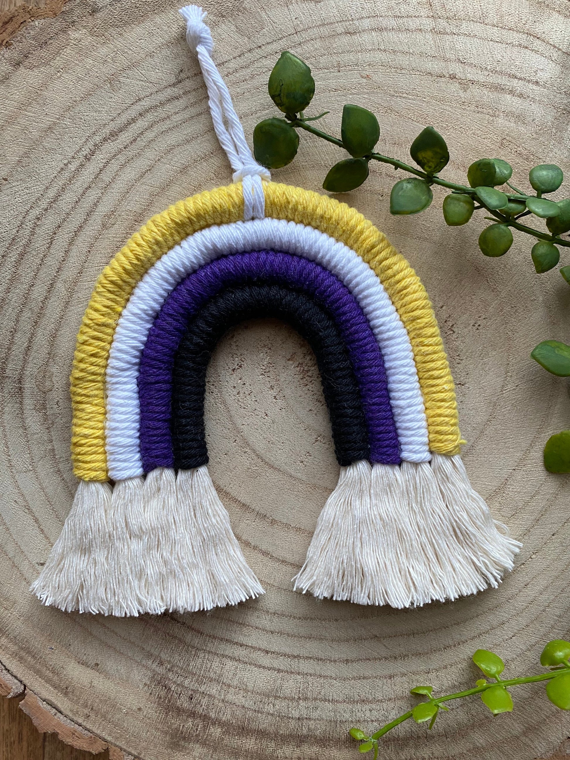 A small macrame rainbow made with yellow white purple and black yarns to represent the colours of the non binary pride flag, with a white hanging loop, placed on a wooden surface with green plant vines.