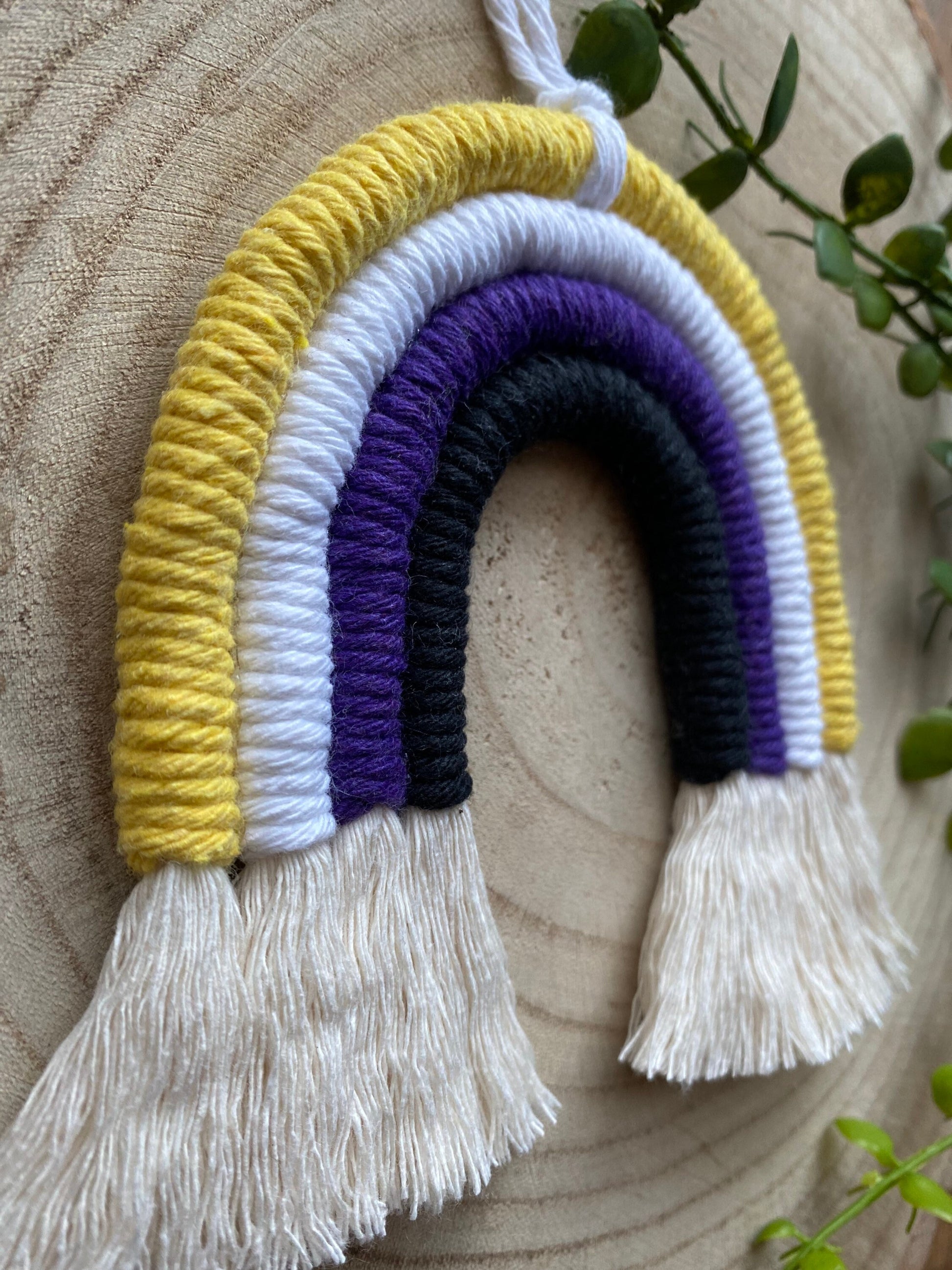 A close up side angle of a small macrame rainbow made with yellow white purple and black yarns to represent the colours of the non binary pride flag, with neatly brushed out tassels at the bottom.