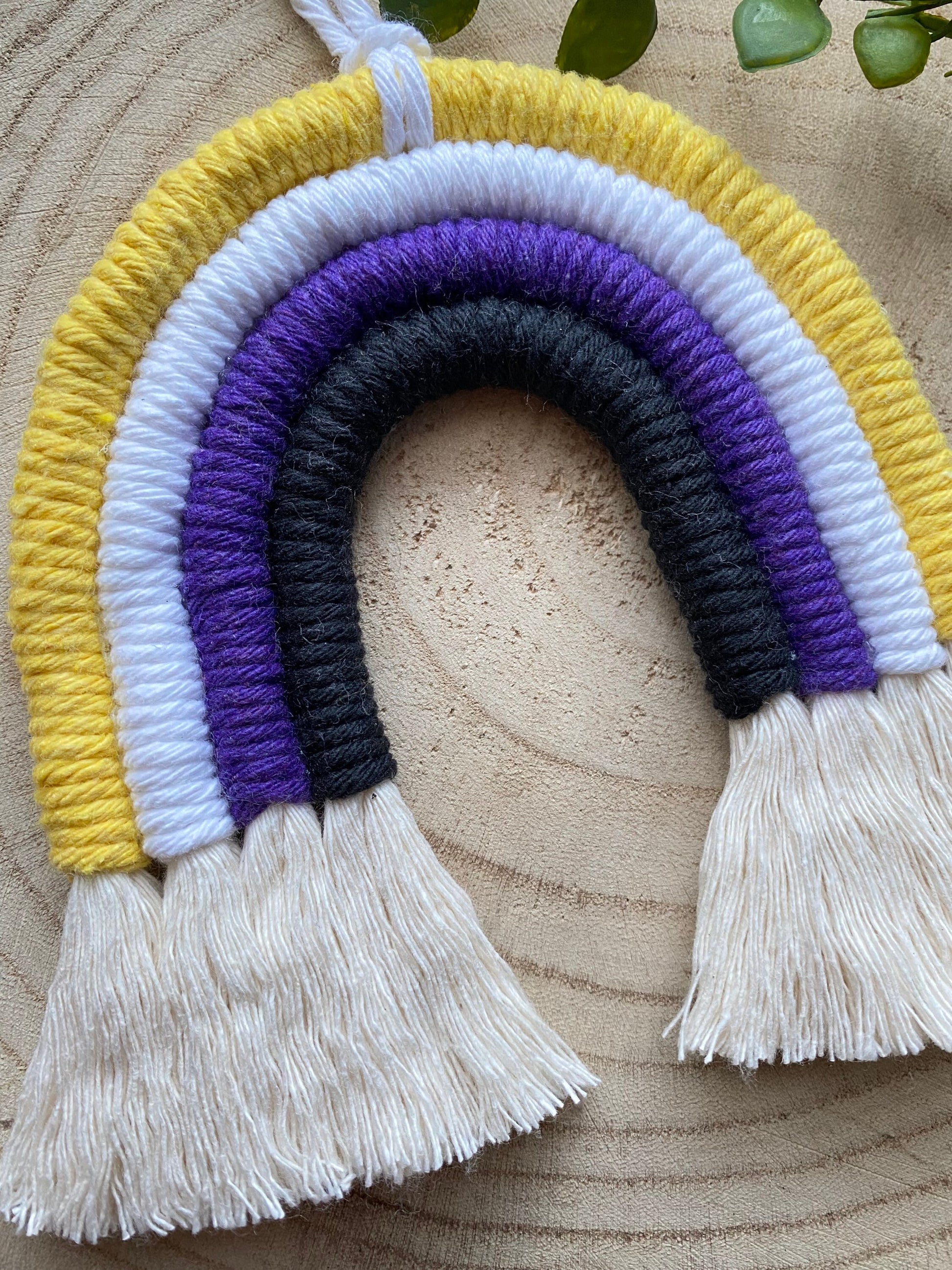 Close up of a small macrame rainbow made with yellow white purple and black yarns to represent the colours of the non binary pride flag, with brushed out tassels at the bottom.