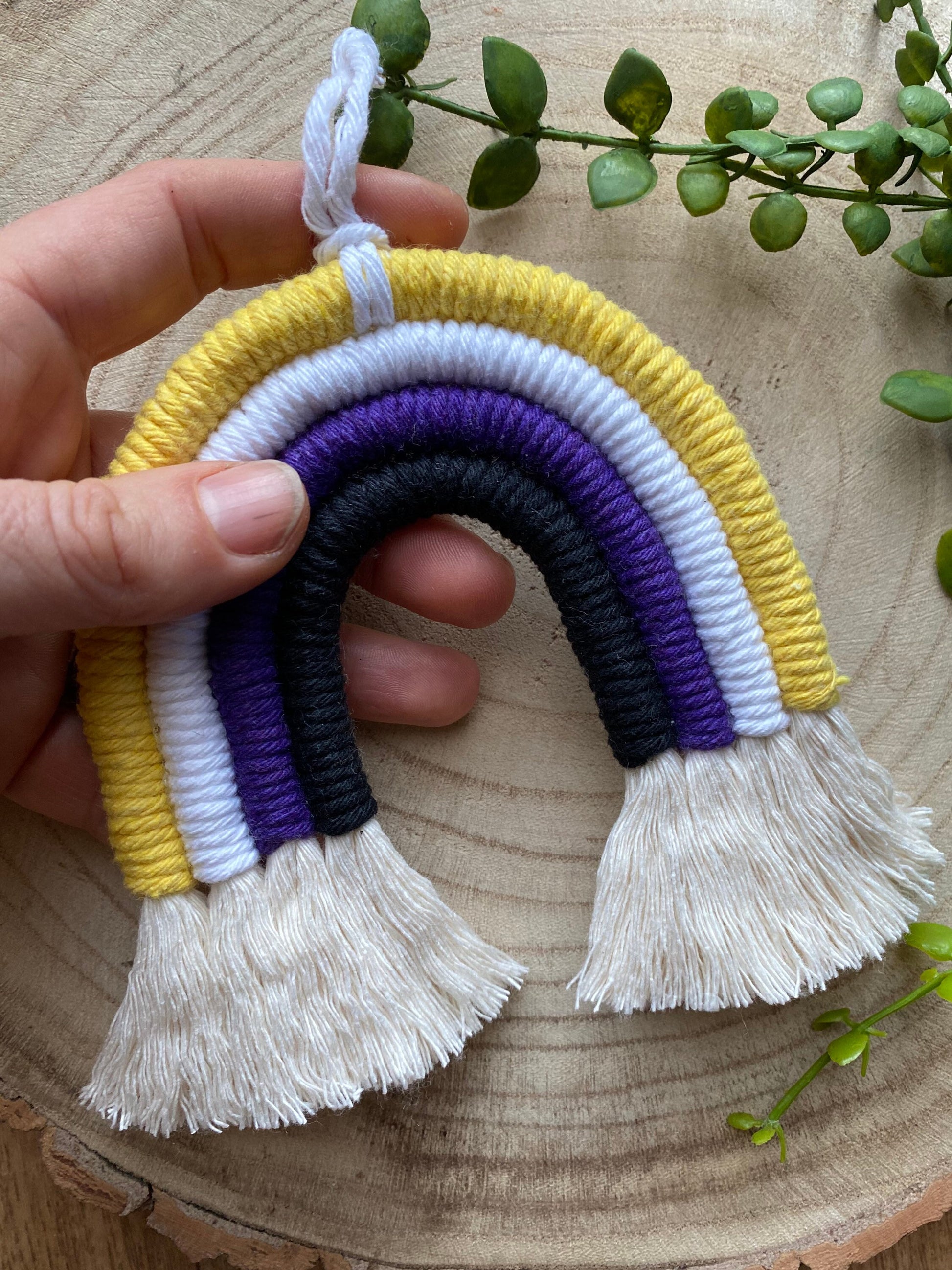 A hand holding a small macrame rainbow made with yellow white purple and black yarns to represent the colours of the non binary pride flag, with fluffy tassels at the bottom.