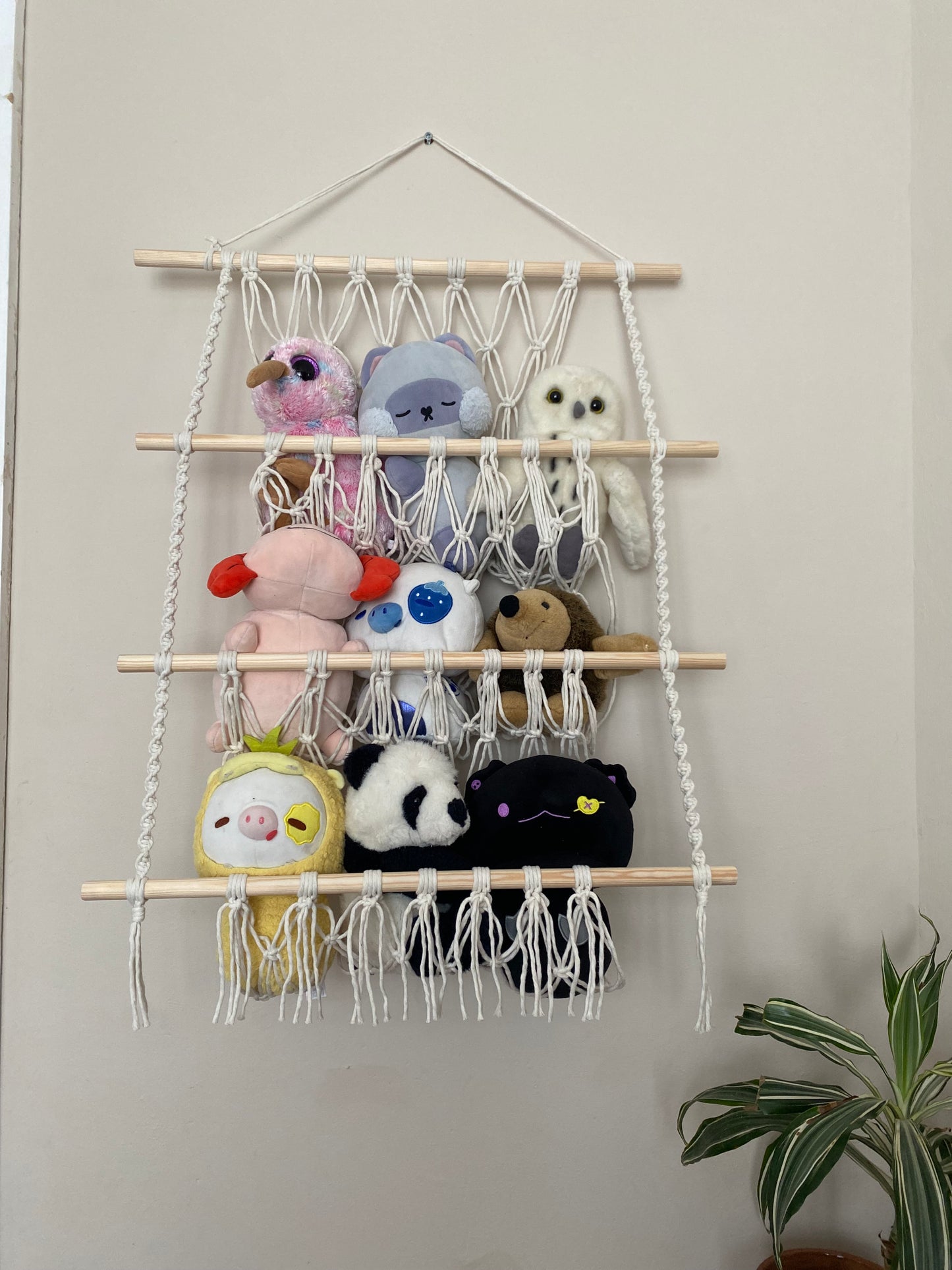 A macrame toy hammock with three layers, made with natural yarn and wooden dowels, hanging against a beige wall, filled with soft toys and teddies.
