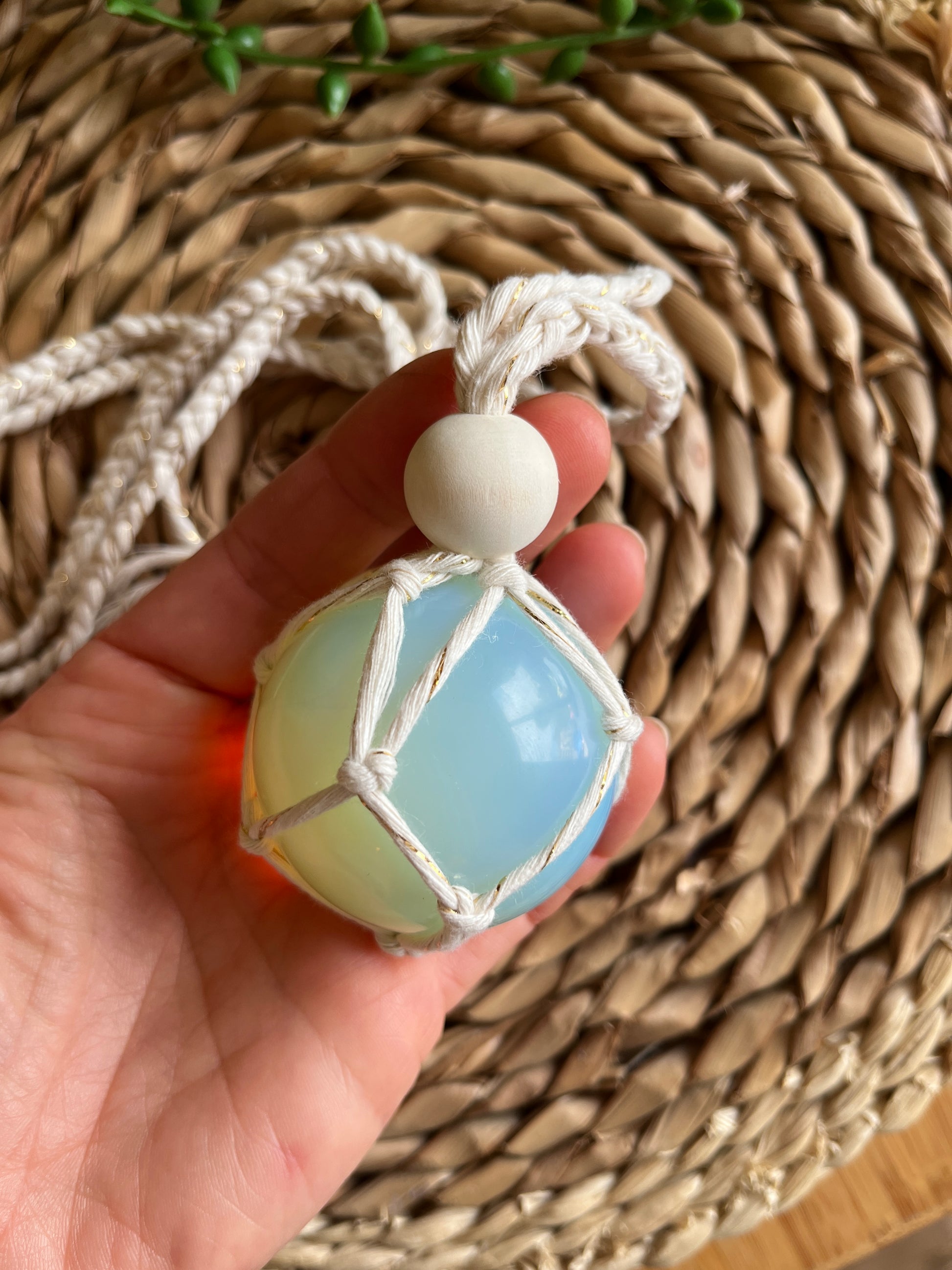 A hand holding a handmade macrame opaline sphere hanging sun catcher, made with eco friendly natural recycled cotton yarn. There is a wicker mat in the background, there is a plain wooden bead above the crystal.