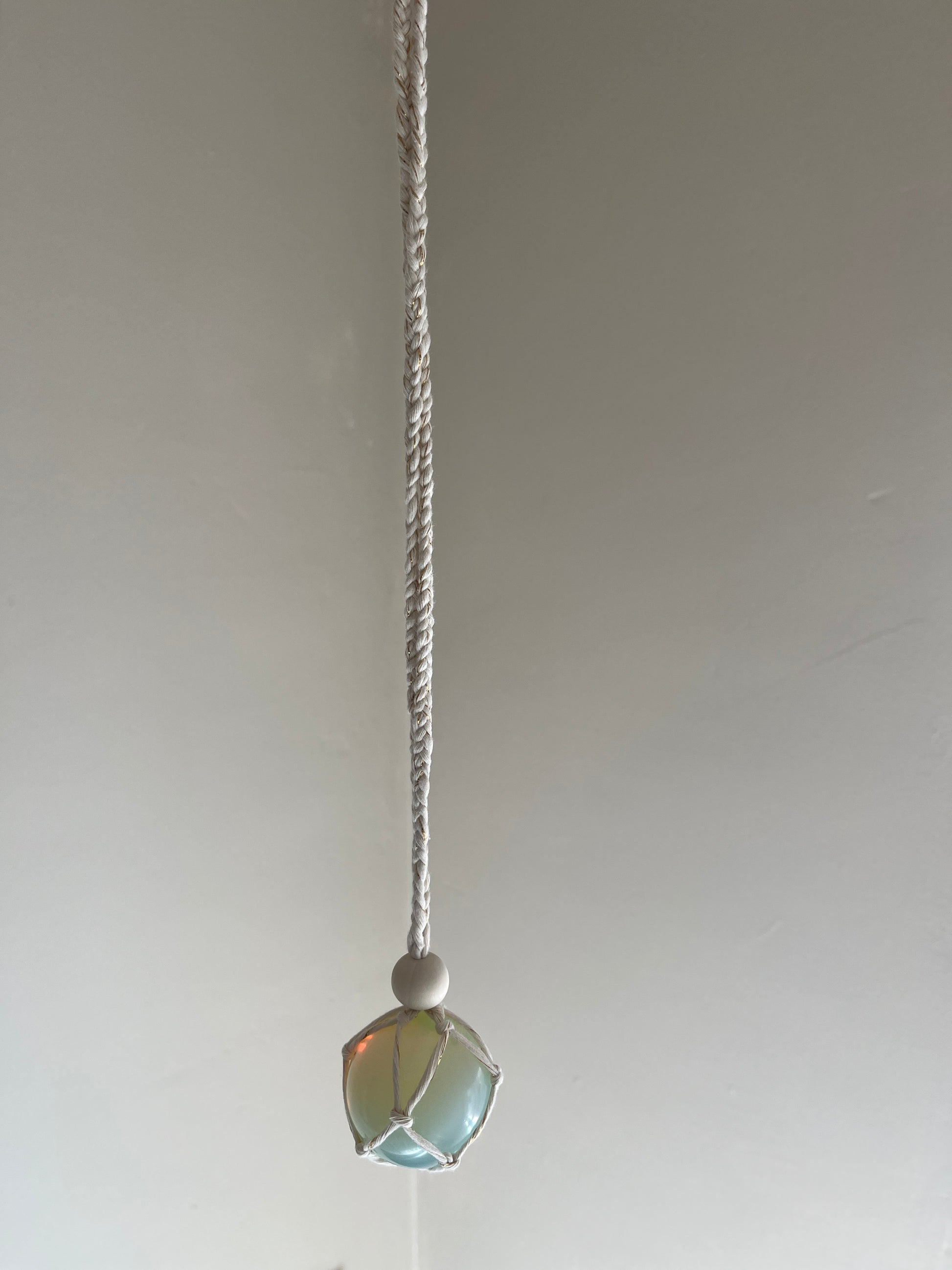 Handmade macrame opaline sphere hanging sun catcher, made with eco friendly natural recycled cotton yarn. The hanger is shown in front of a plain white wall, there is a plain wooden bead above the crystal.