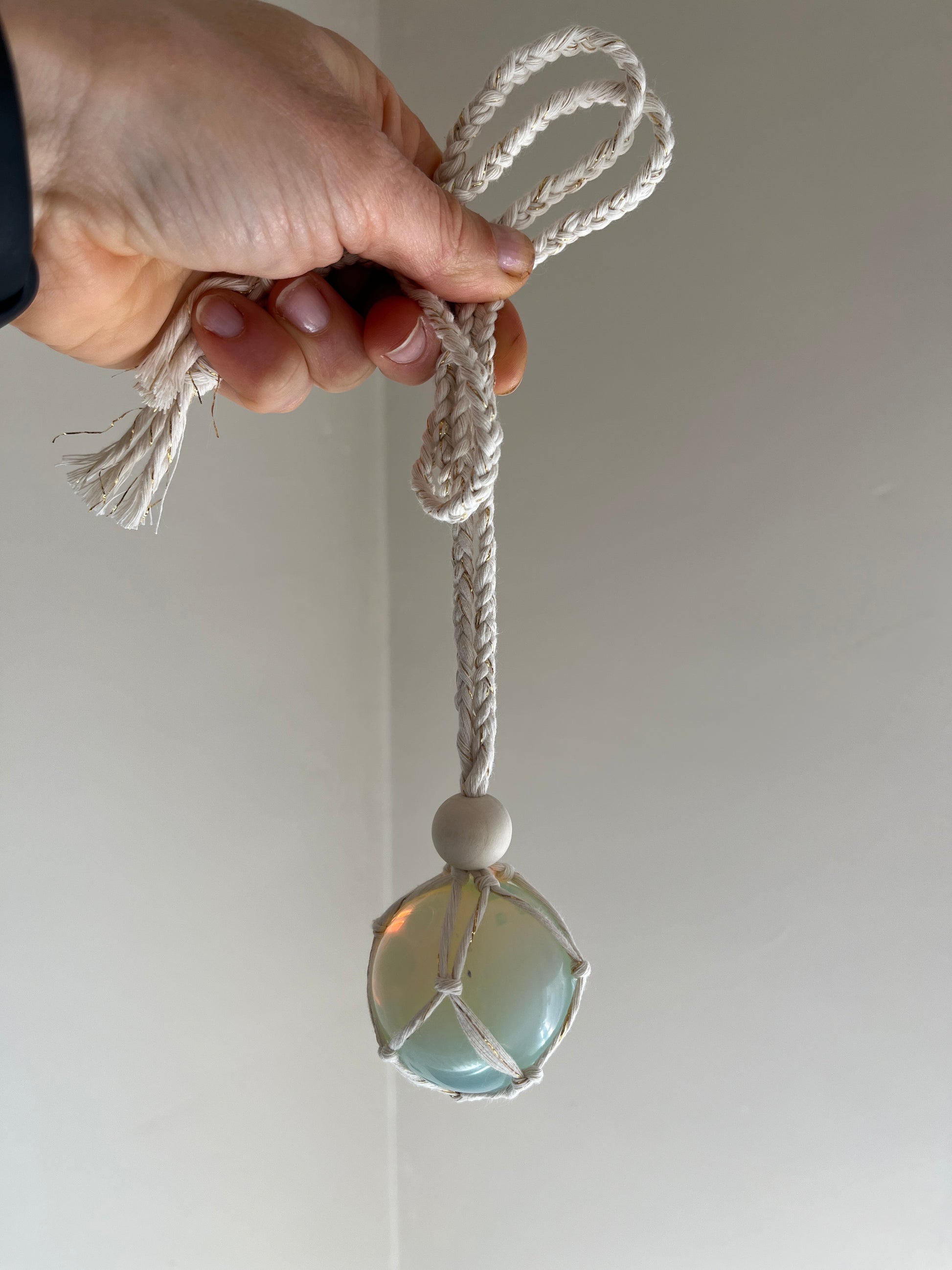 Handmade macrame opaline sphere hanging sun catcher, made with eco friendly natural recycled cotton yarn. The hanger is shown being held in front of a plain white wall, there is a plain wooden bead above the crystal.