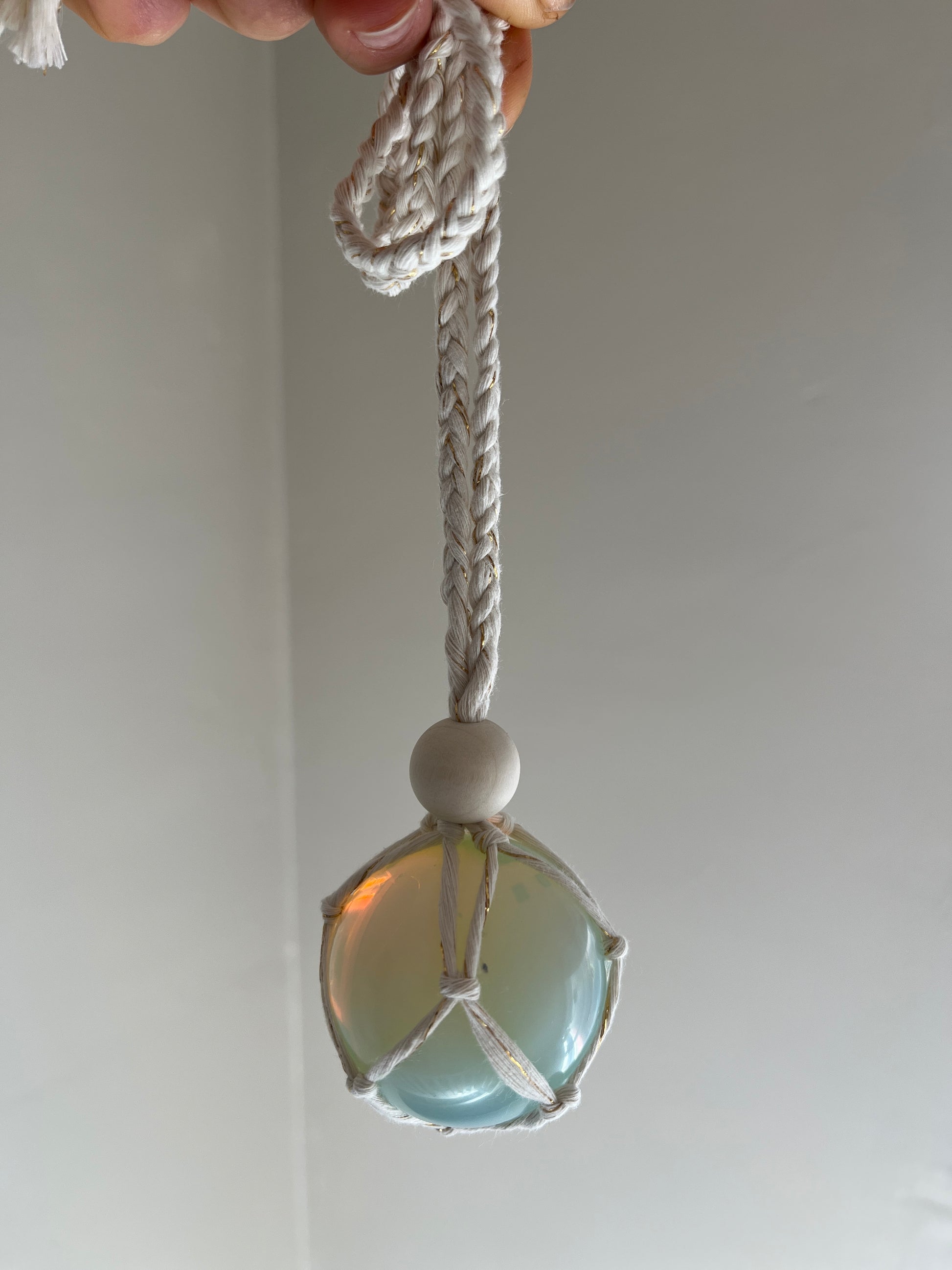 Handmade macrame opaline sphere hanging sun catcher, made with eco friendly natural recycled cotton yarn. The sphere is shown held by a hand,  hanging in front of a plain white wall, there is a plain wooden bead above the crystal.