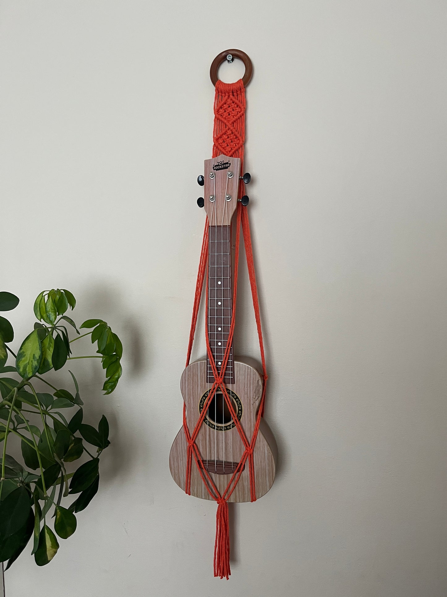 A light brown ukulele sits inside of a handmade macrame wall hanging ukulele hanger, which is made with orange recycled cotton, and a repurposed wooden hanging ring. A large green leafy plant is to the left of the image.
