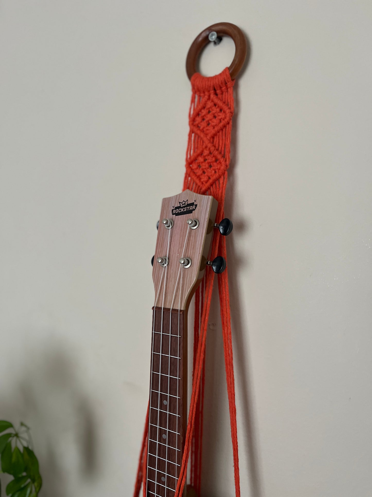 A light brown ukulele sits inside of a handmade macrame wall hanging ukulele hanger, which is made with orange recycled cotton, and a repurposed wooden hanging ring.