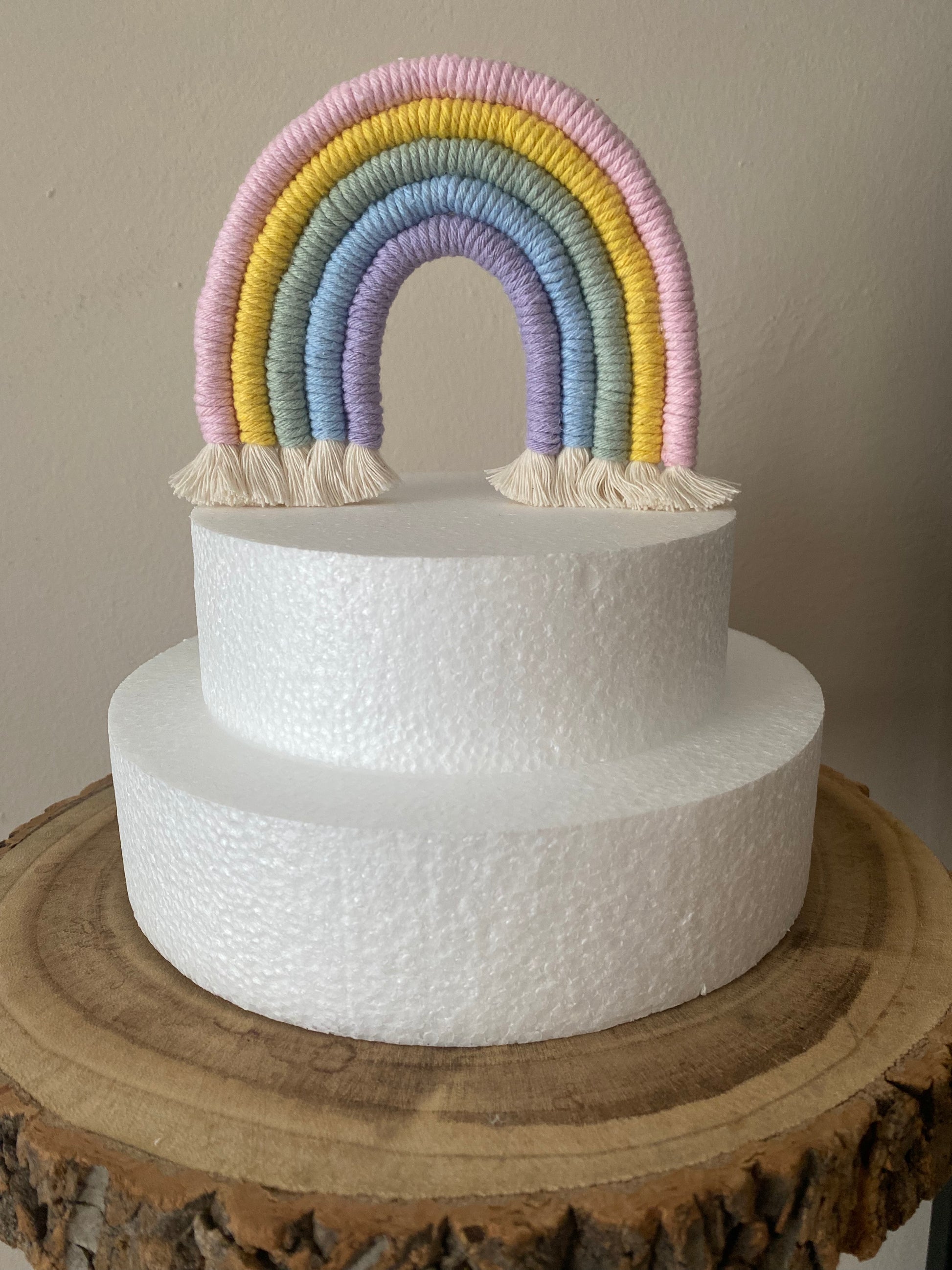 Pastel rainbow macramé cake topper on a white polystyrene cake, atop a wooden board.