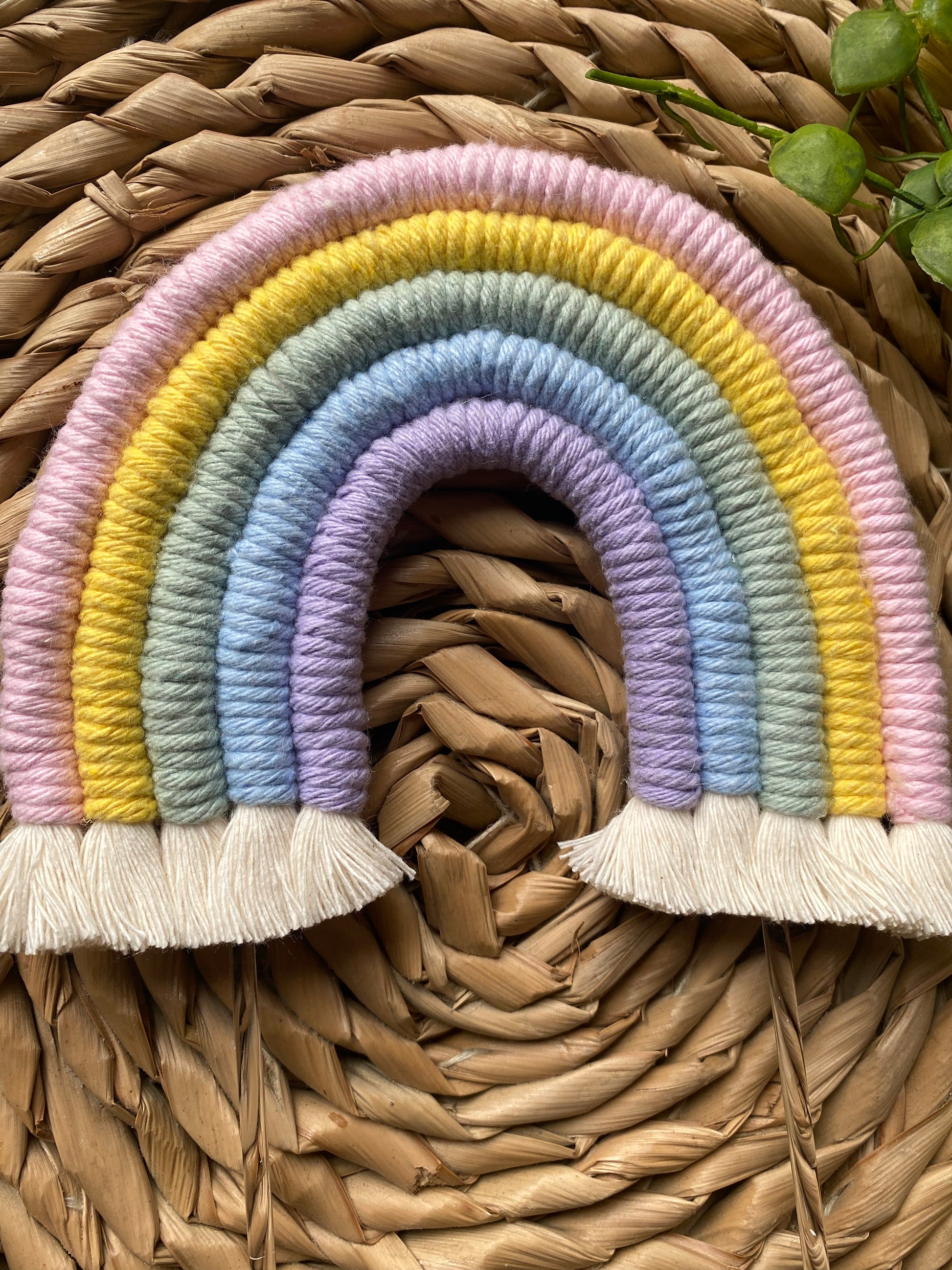 Handmade pastel rainbow macramé cake topper with long clear acrylic sticks, laid on a round wicker mat.